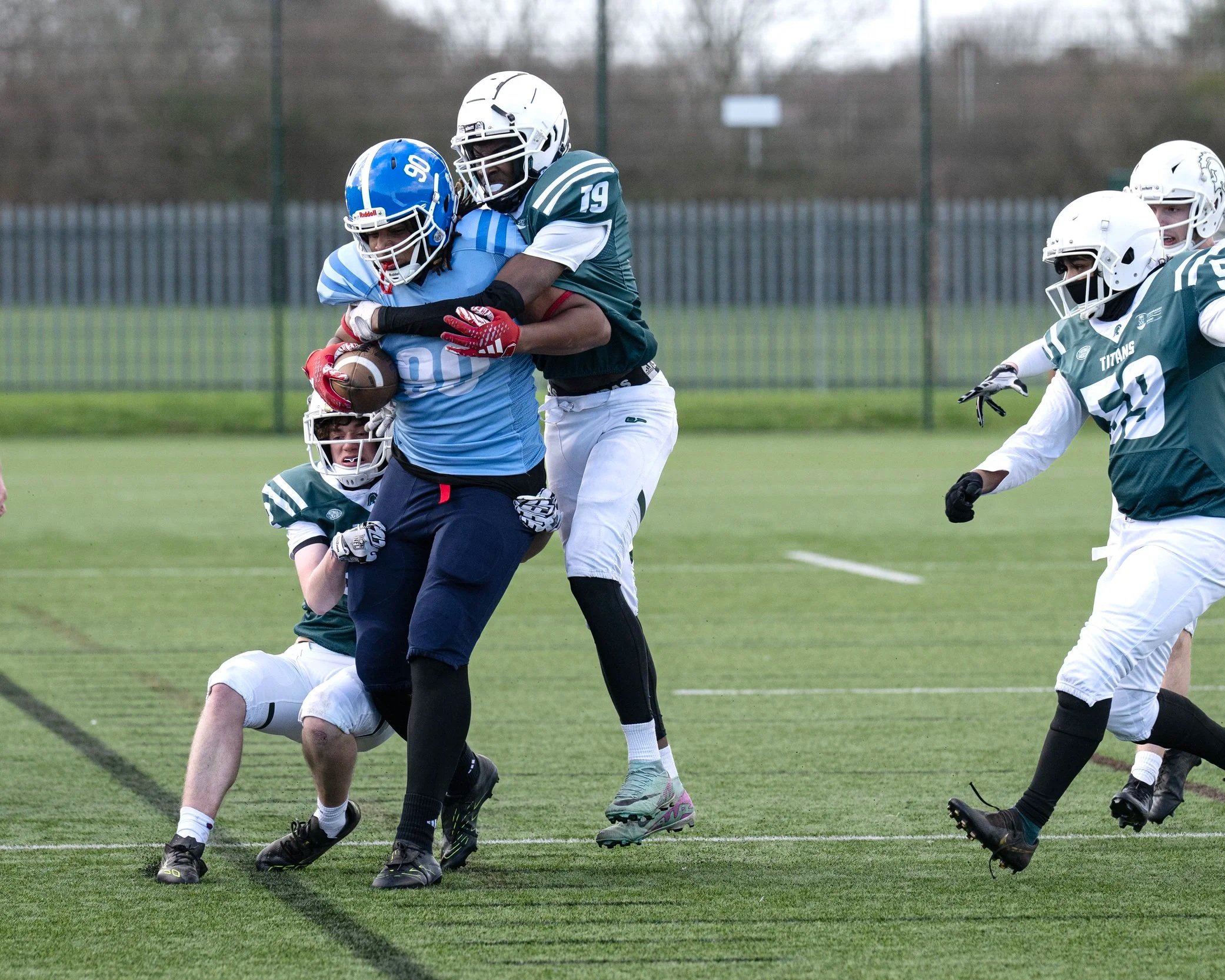 American football game with players tackling and running on a green field with a fence and trees in the background.