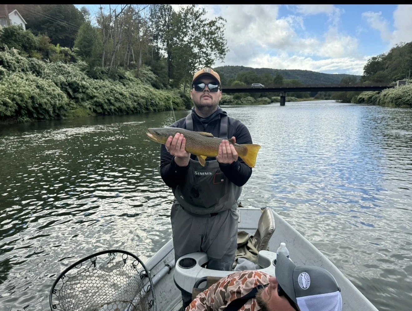 Man holding a large fish he caught on a boat in a river with a bridge in the background, surrounded by green trees.
