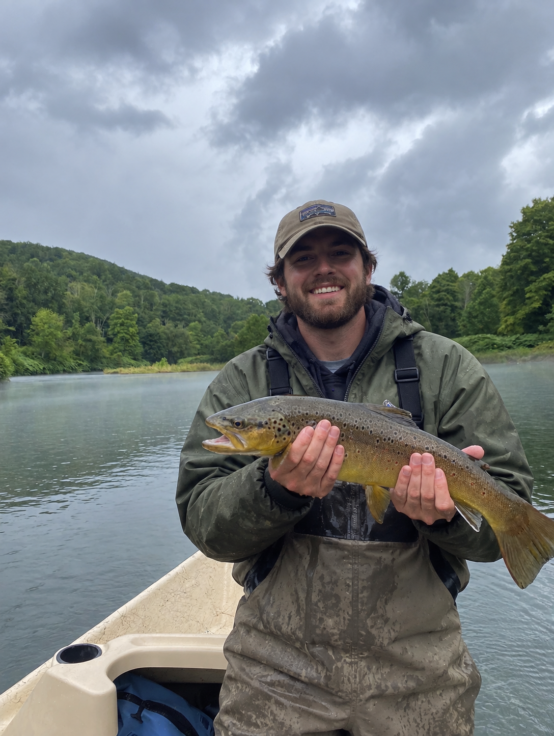 A man in outdoor clothing holding a large fish on a boat, with a river and green hills in the background on a cloudy day.