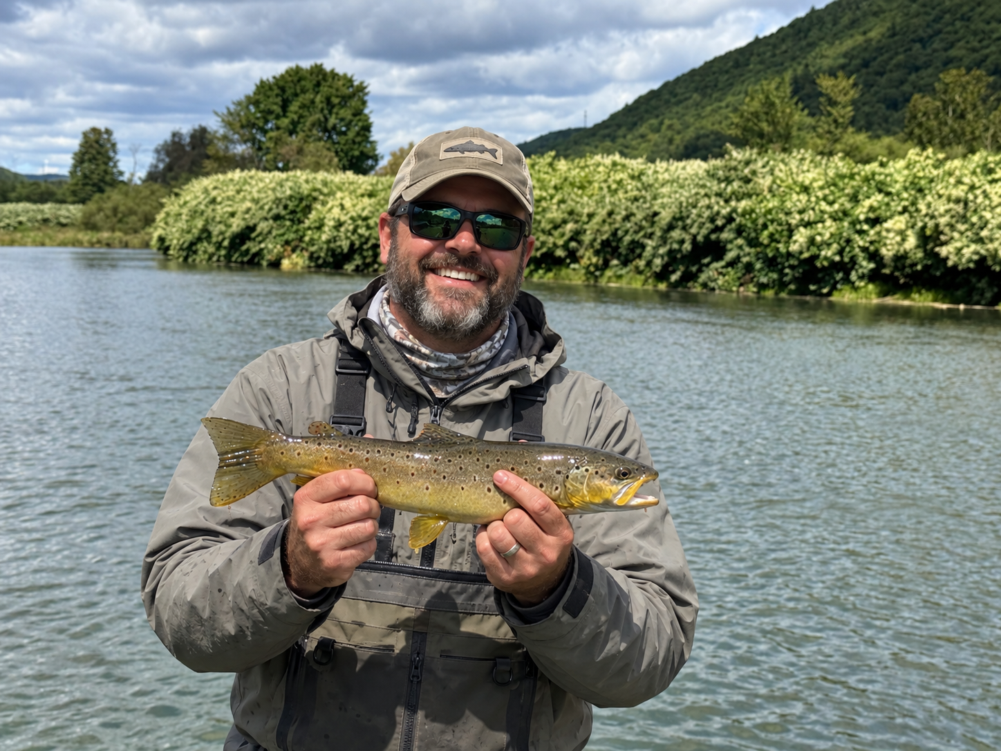 Man with sunglasses smiling and holding a fish by the water, with green trees and a hill in the background.
