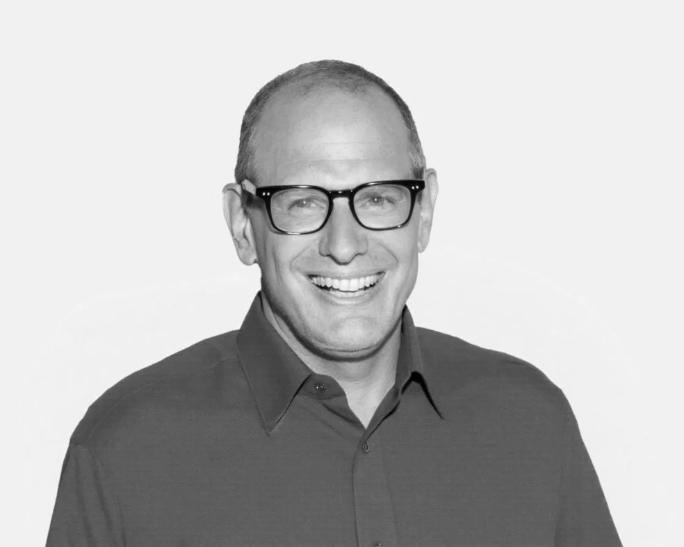 A black-and-white portrait of a man smiling, wearing glasses and a button-up shirt.