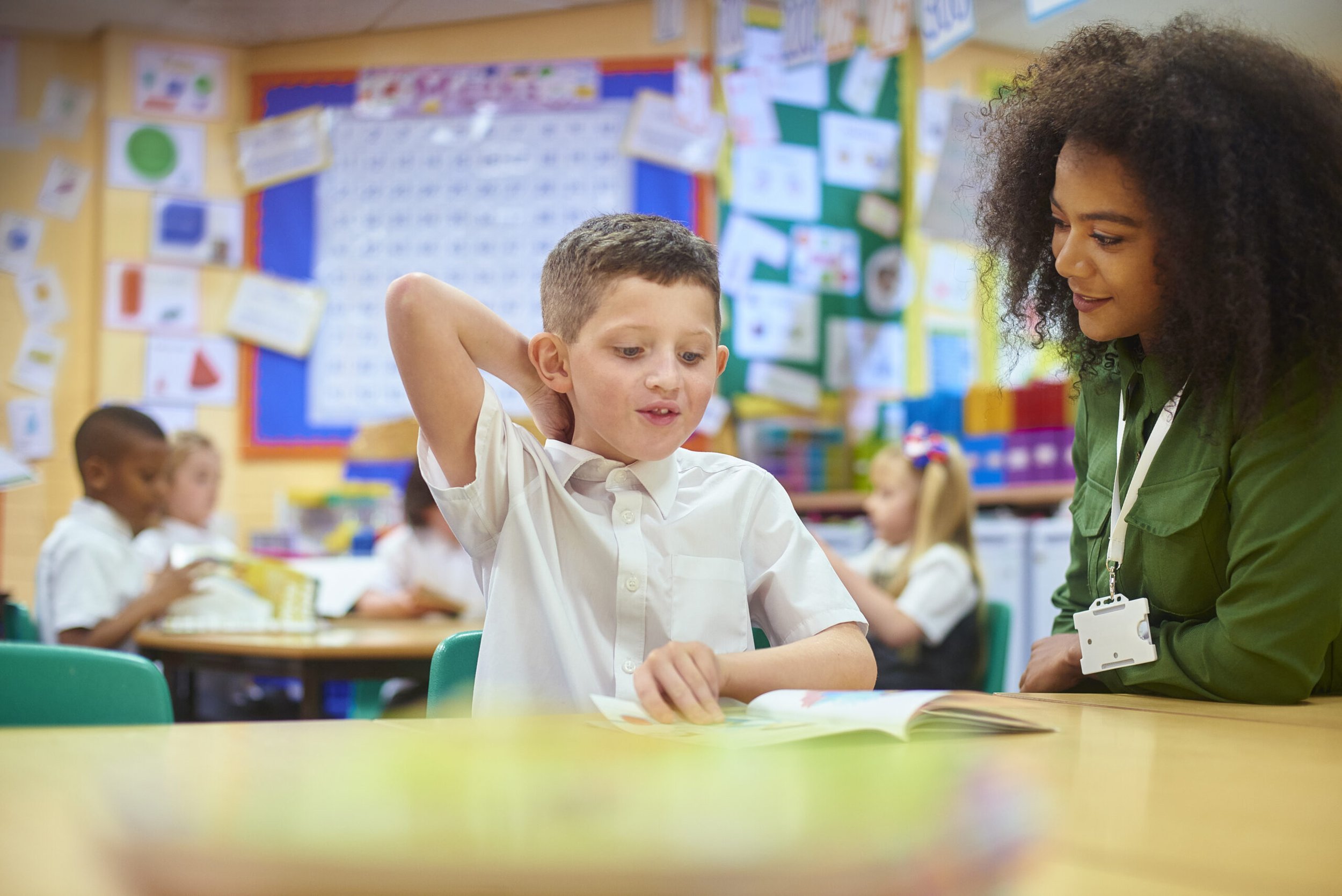 A boy in a white shirt shows a woman, probably a teacher, a picture book in a colorful classroom with other children in the background.
