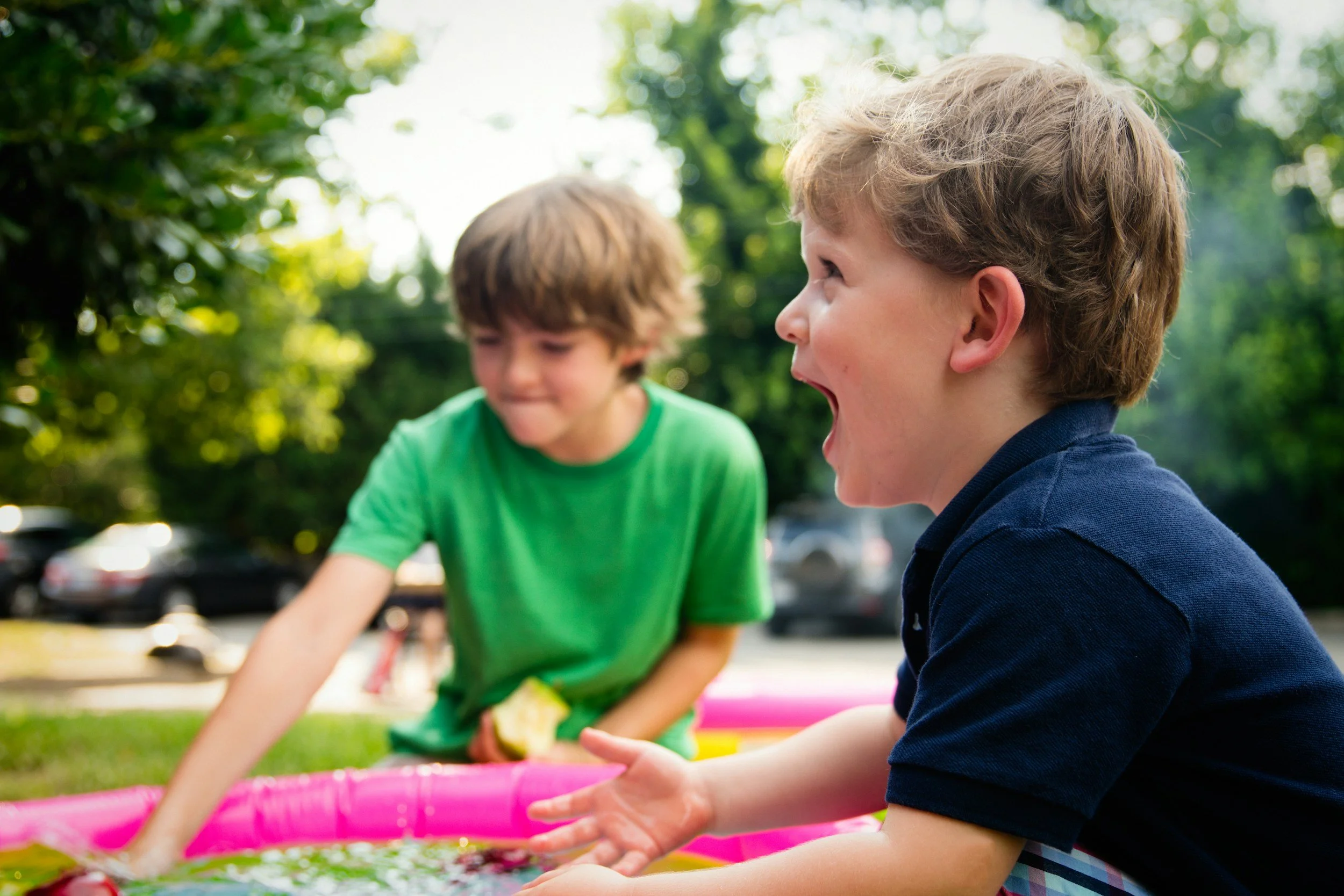 Two young boys playing and splashing water in a pink inflatable pool outdoors on a sunny day with green trees in the background.