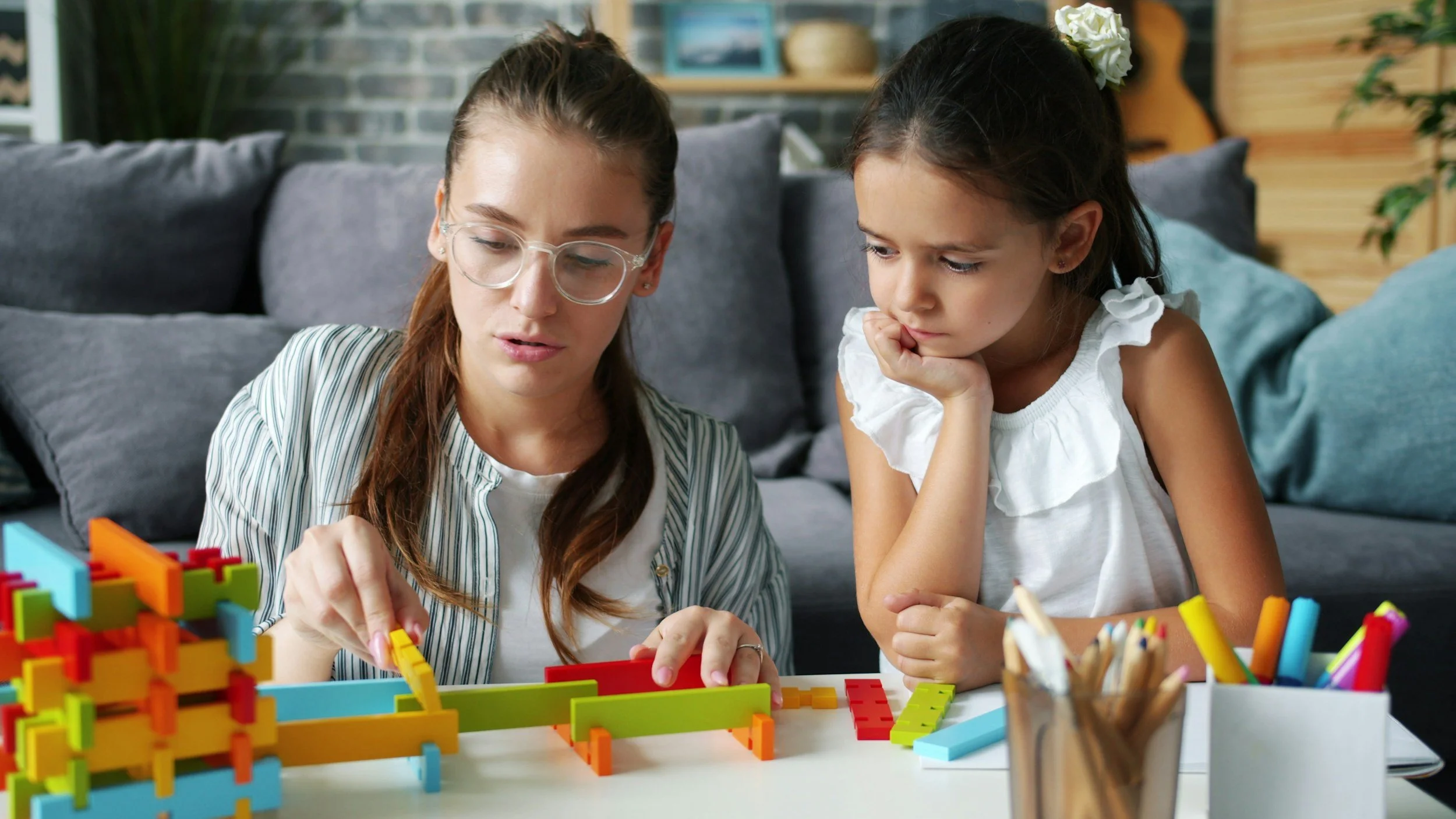 An adult woman and a young girl are playing with colorful building blocks on a table in a living room with gray couch and wooden shelves in the background.