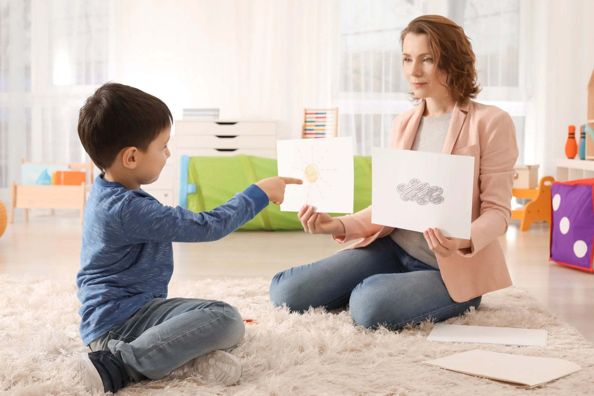 A woman sitting on a beige rug with a young boy, holding up drawings of a sun and a cloud while the boy points to the sun drawing, in a brightly lit room with colorful furniture and toys.