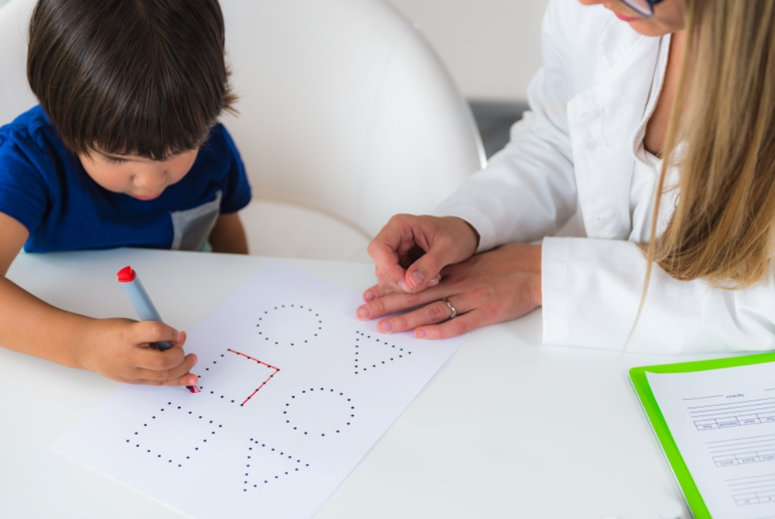 A young boy and a woman are sitting at a table, playing connect-the-dots on a sheet of paper; the boy is using a red marker and the woman is watching closely.