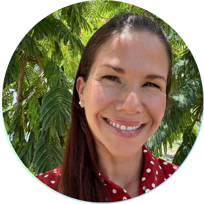 A woman smiling outdoors with lush green foliage in the background, wearing a red shirt with white polka dots.