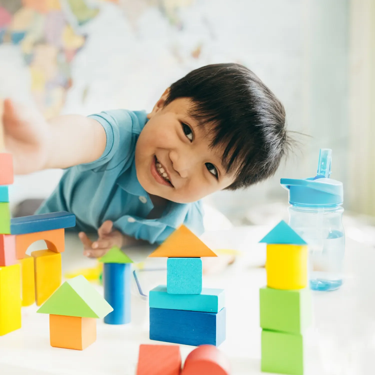 A young boy with dark hair smiling and reaching toward colorful wooden building blocks on a table.