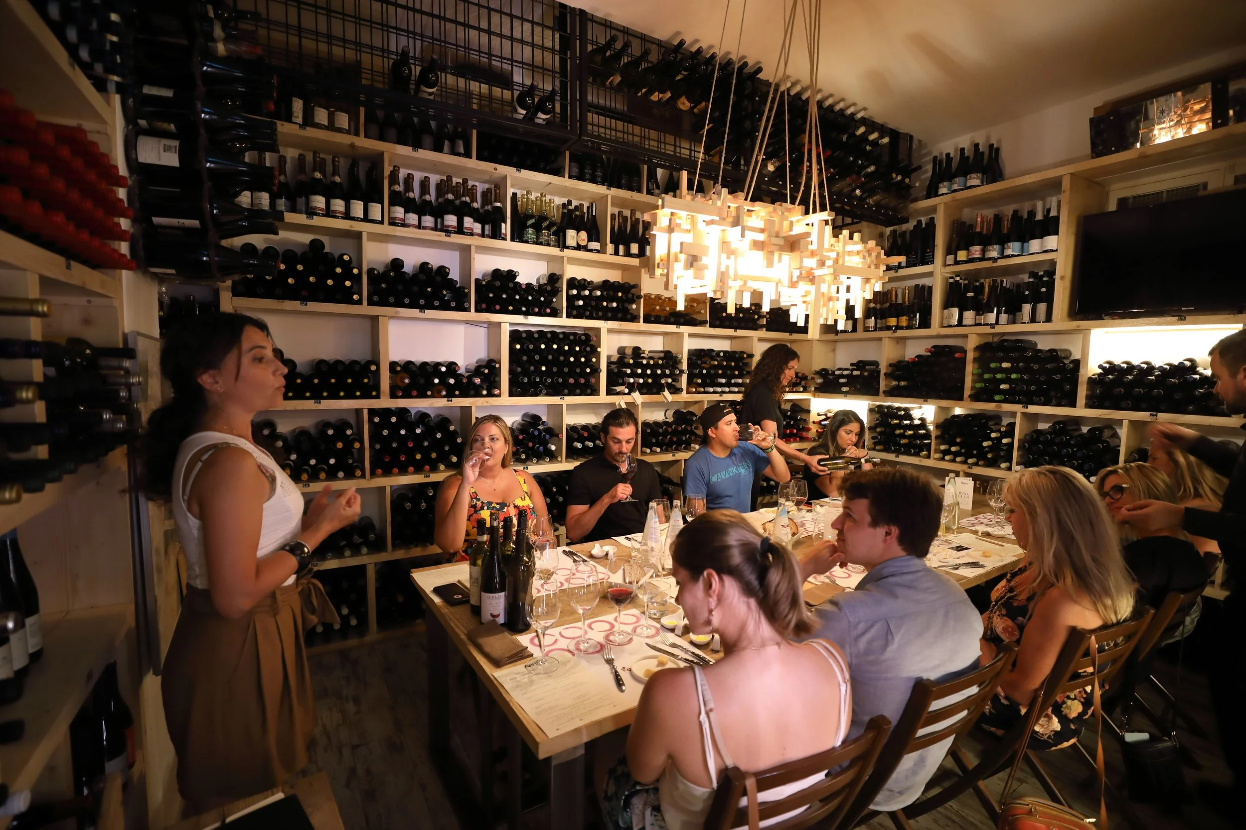 A group of people seated at a long table inside a wine cellar restaurant, tasting and drinking wine, with shelves of wine bottles on the walls, and a woman standing at the end of the table.