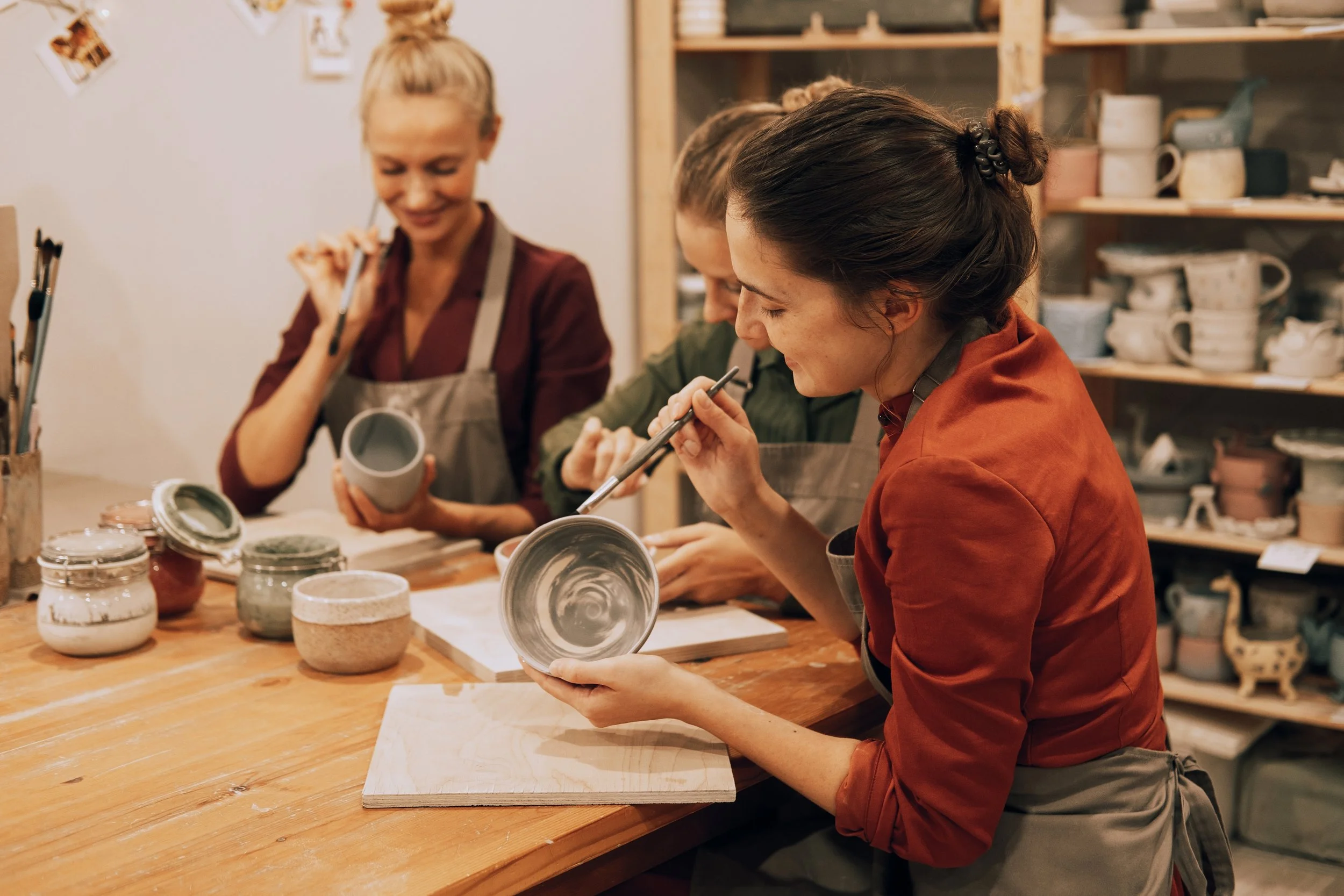 A group of women is working on pottery at a pottery studio, painting and examining glazed ceramic bowls and mugs.
