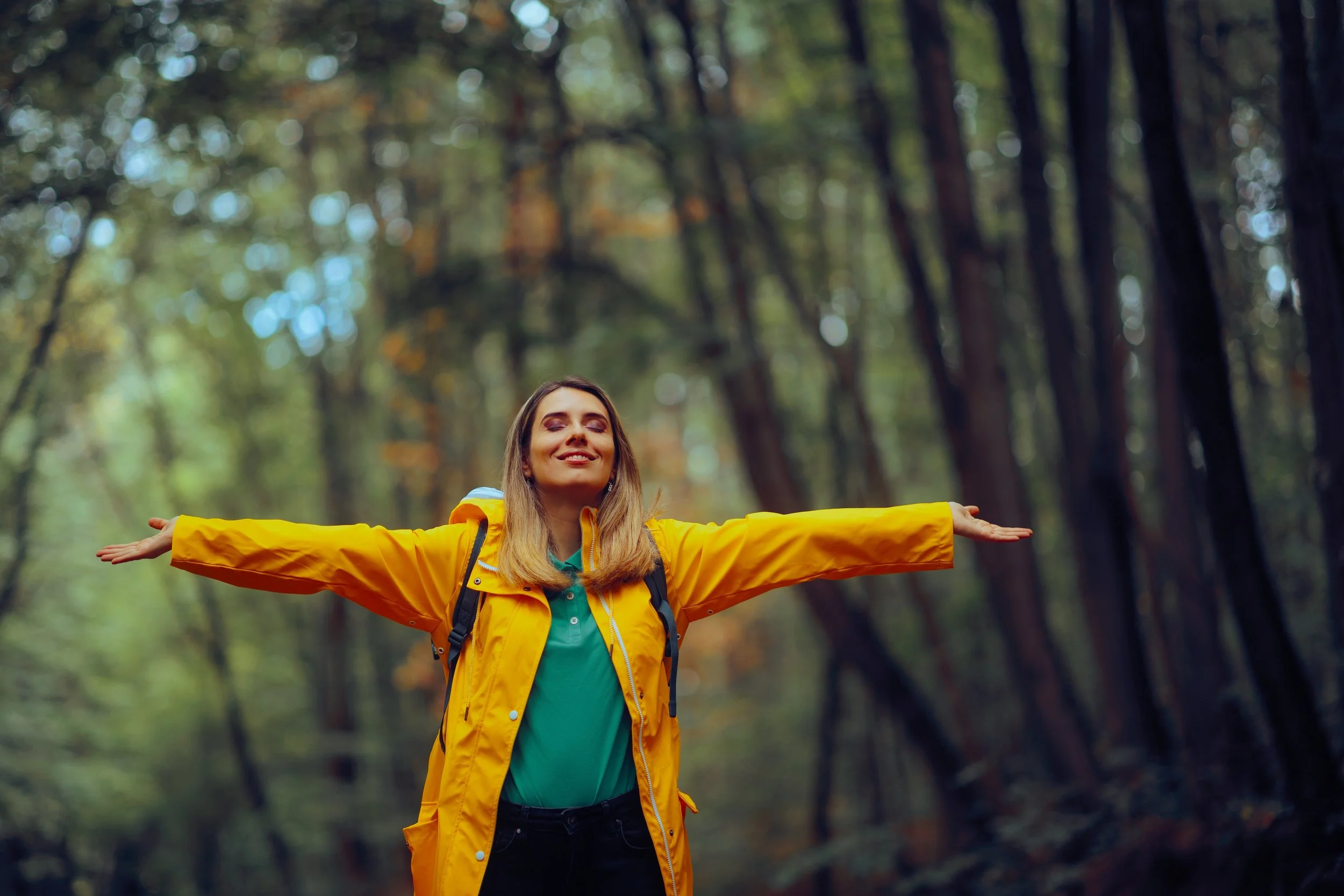 Woman enjoying outdoors in the forest with arms outstretched, wearing a yellow jacket and backpack.