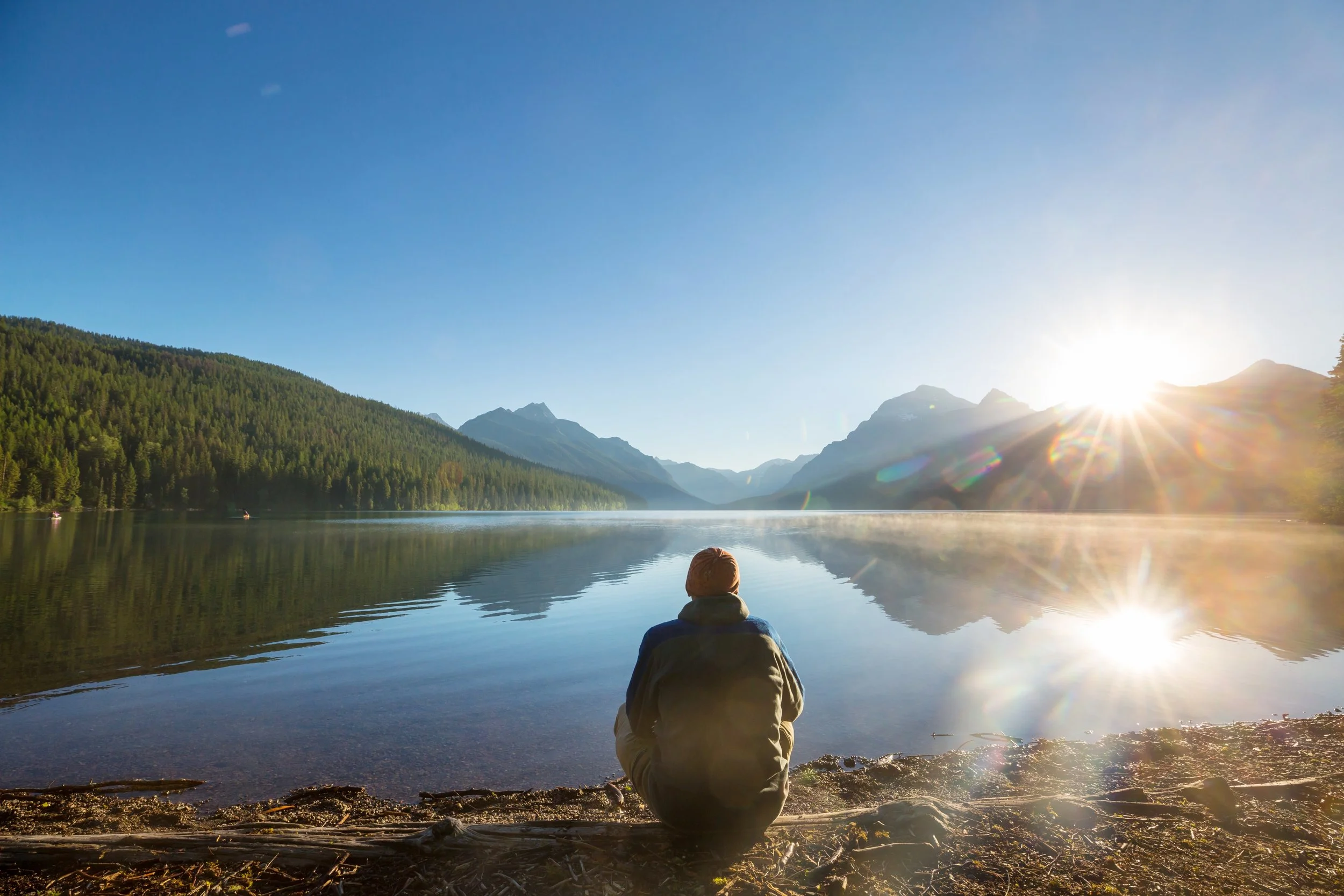 A person sitting on the shore of a calm lake at sunrise, surrounded by mountains and forest, with sunlight reflecting on the water.