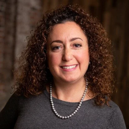A woman with curly hair, wearing a dark top and a pearl necklace, smiling in an indoor setting.