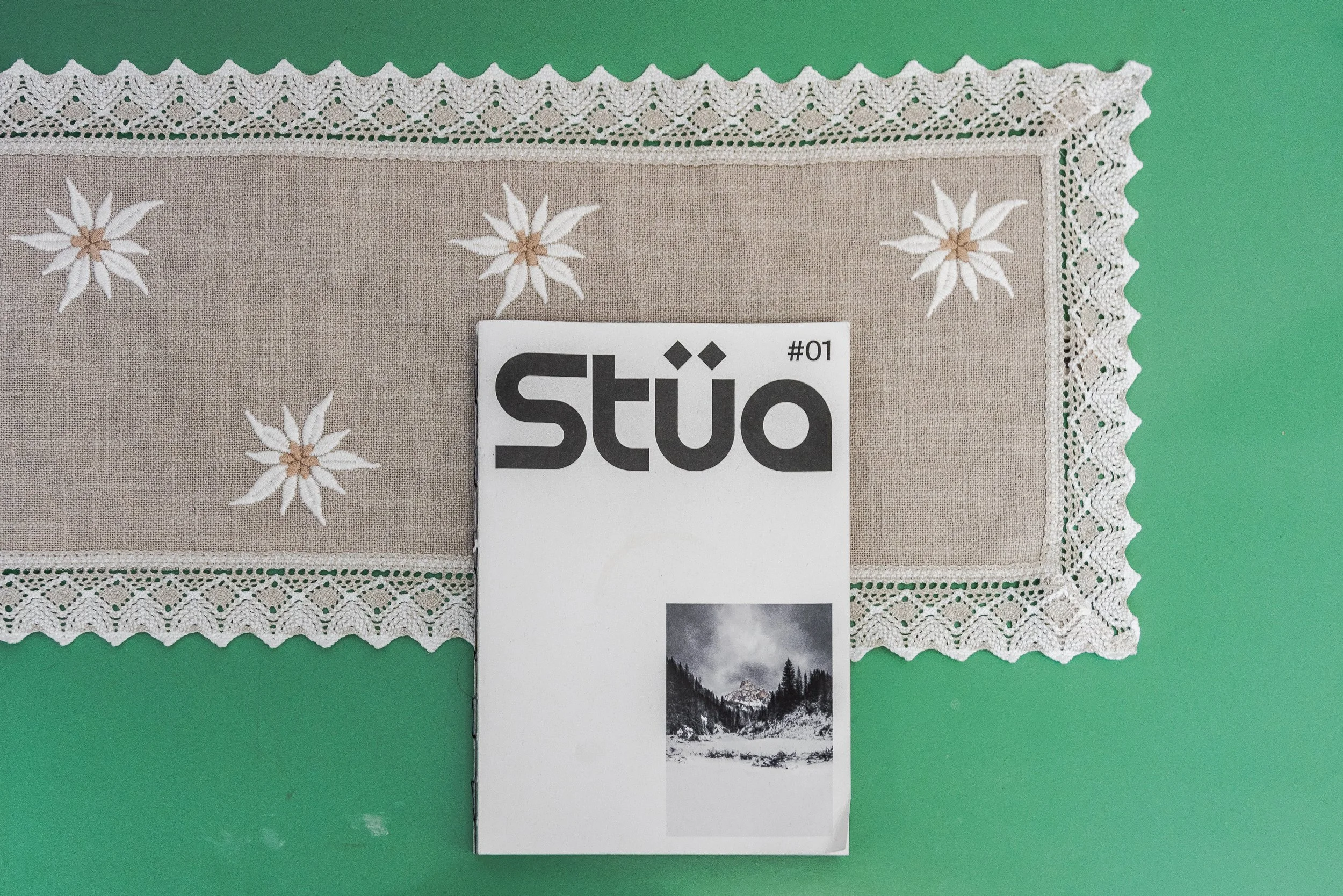Un tovagliolo di pizzo bianco con decorazioni di fiore e un'etichetta con il testo 'Stüb #01' sopra un'immagine di paesaggio montano innevato.