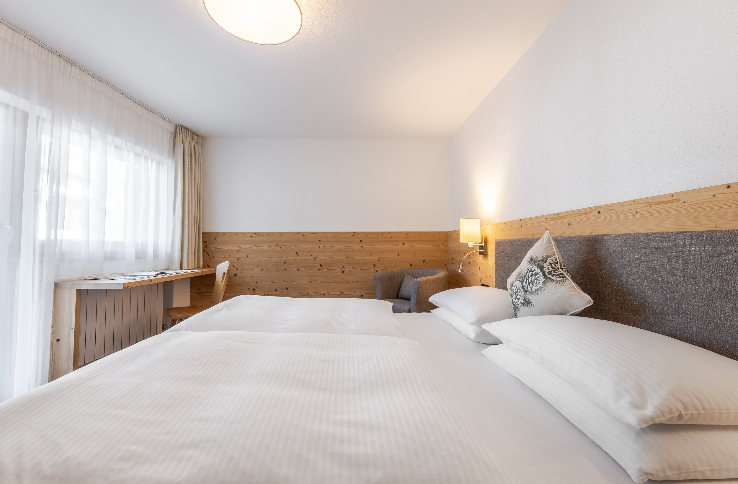 Camera da letto con letto matrimoniale, federe bianche e cuscino decorativo con motivi di pino, parete con pannelli di legno e lampada da parete, grande finestra con tende trasparenti e tende di stoffa, arredamento semplice e luminoso.