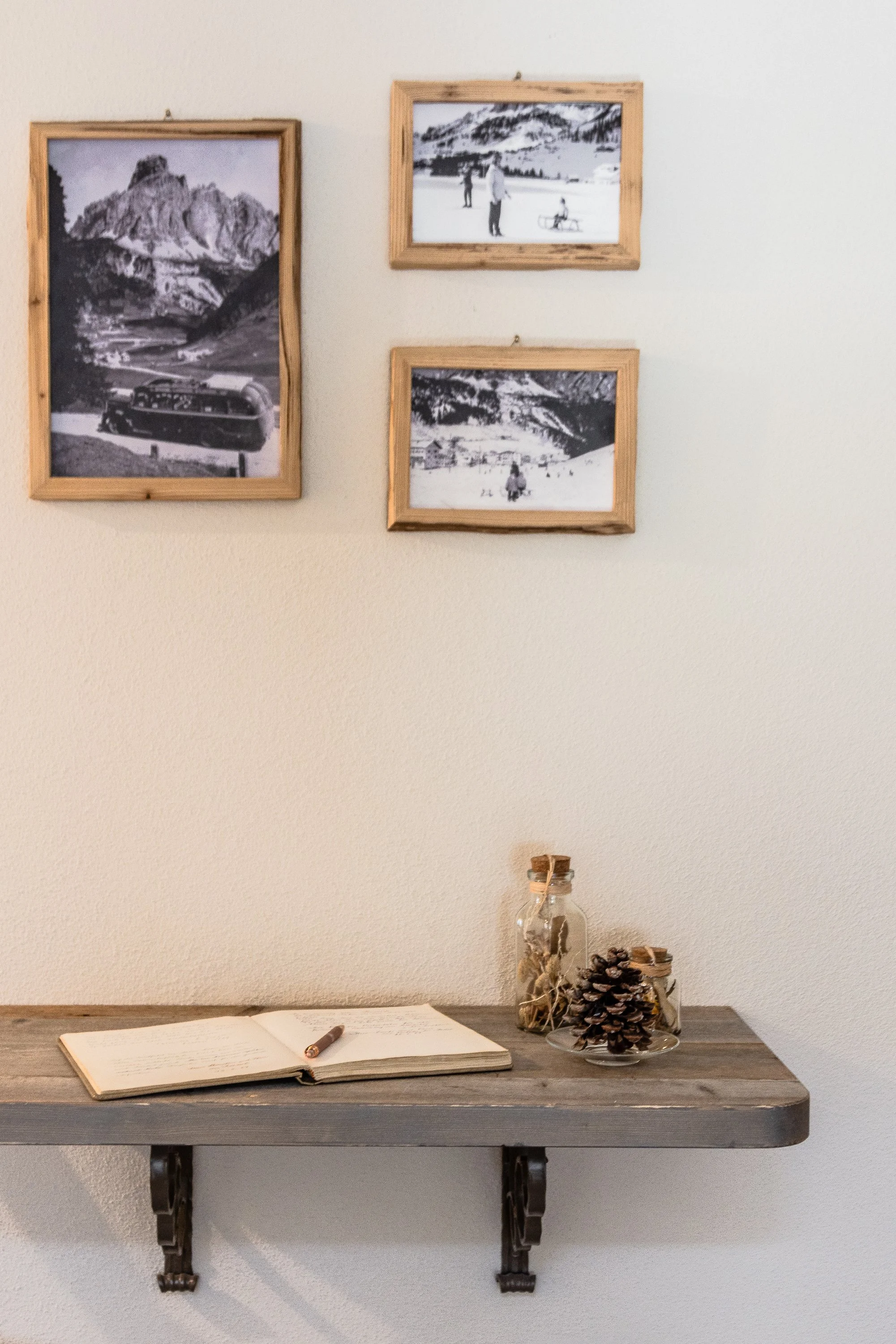 Una scrivania in legno con un libro aperto, una penna e decorazioni di pigne nel vetro, un muro con quattro cornici con foto di paesaggi montani e scene di neve.