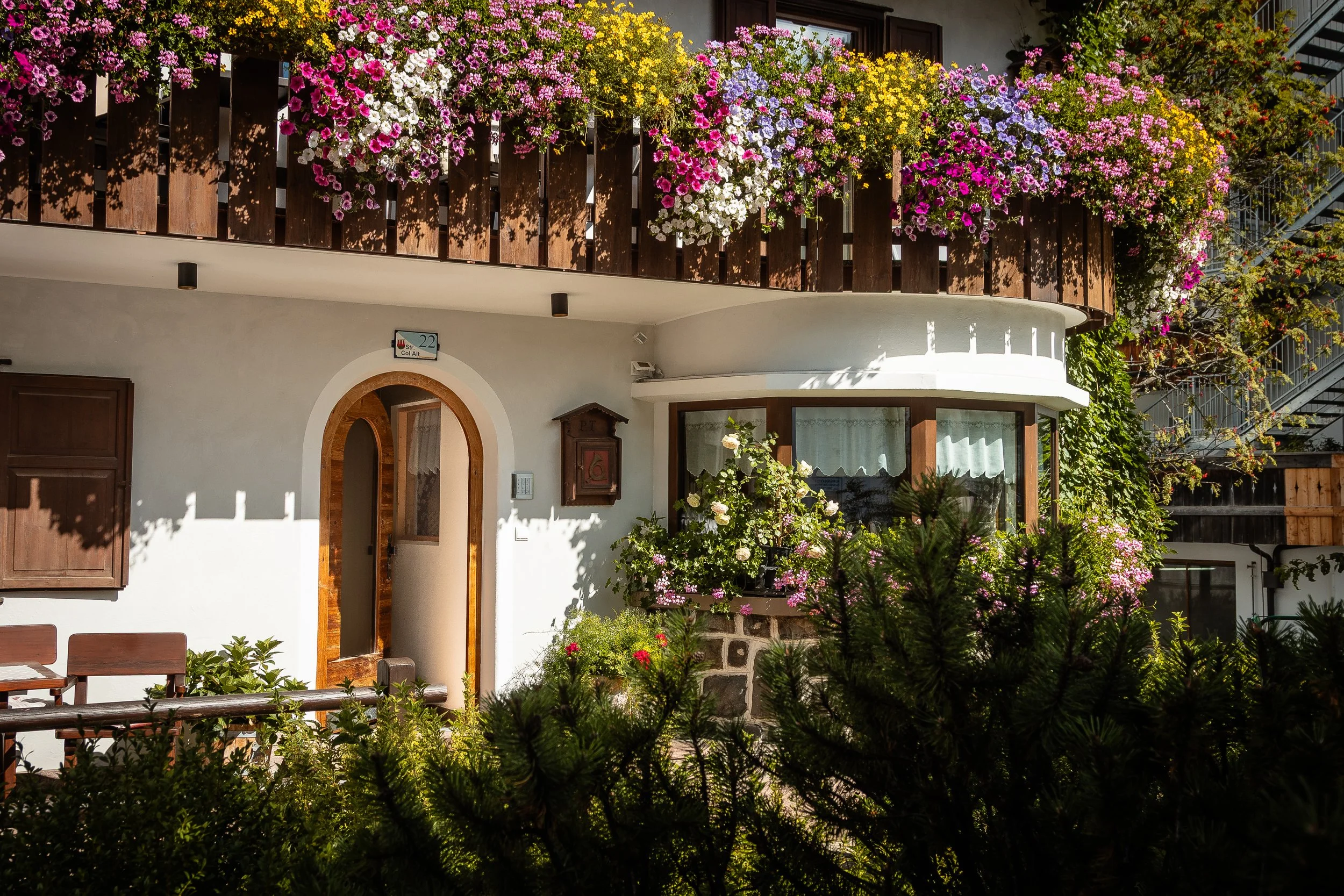Facciata di un edificio con balcone decorato con molte piante e fiori colorati, tra cui petunie, gerani e altri, in un contesto cittadino con secondo piano, ringhiera in legno e finestre.