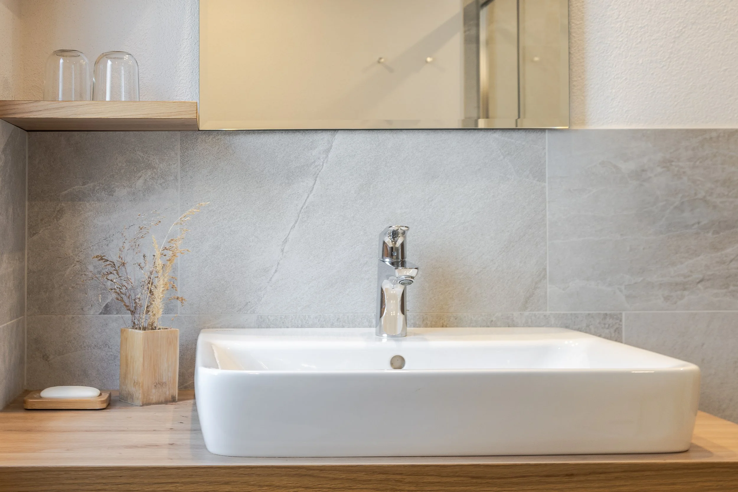 Lavabo bianco con rubinetto in metallo, accanto una pianta secca in un vaso di legno e una candela su un piano in legno, sfondo di pareti con rivestimento di piastrelle grigie.