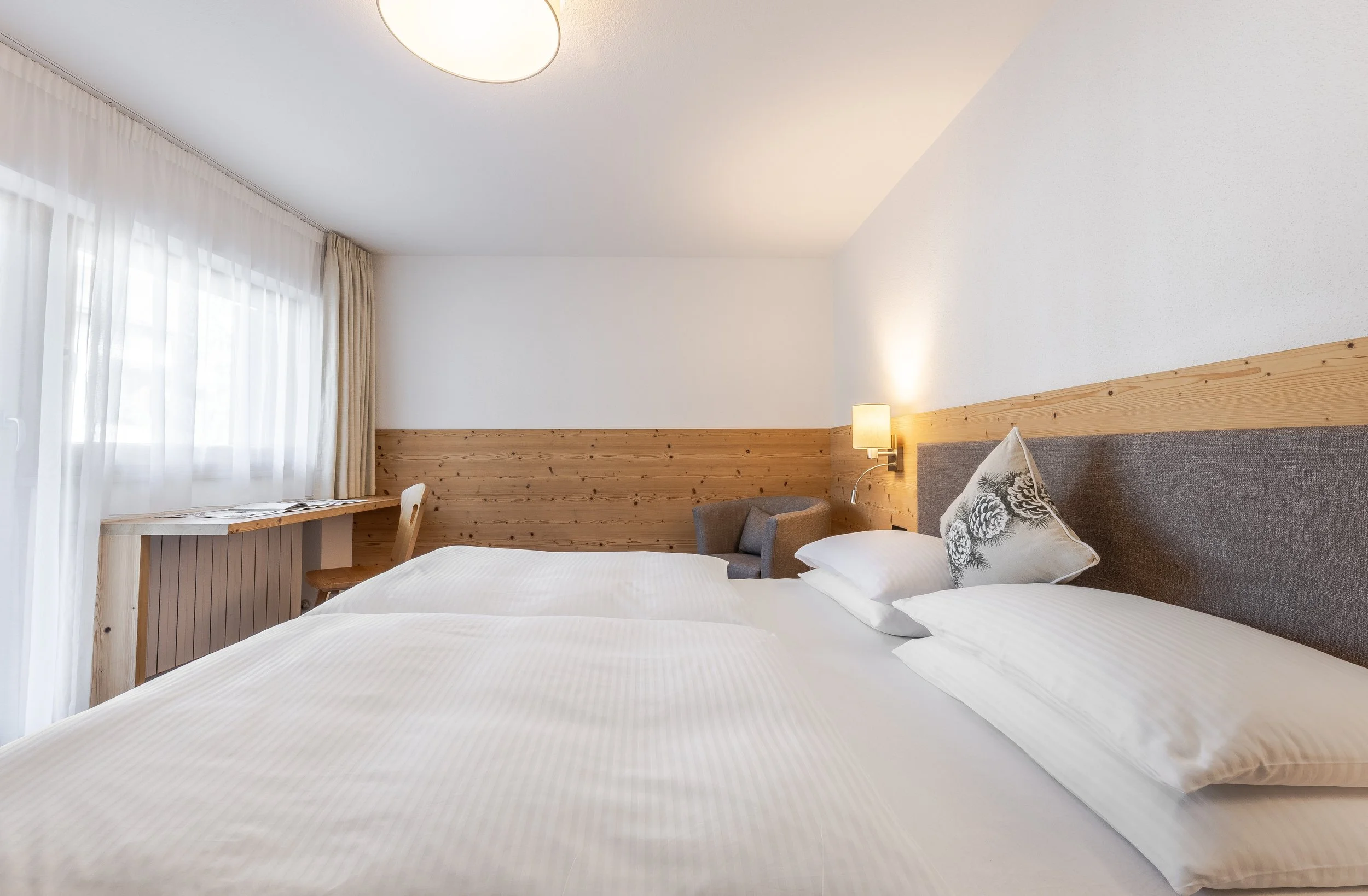 Camera da letto con vers planata, due letti con lenzuola bianche e cuscini, pareti di legno e una lampada accesa, grandi tende bianche che coprono una finestra.