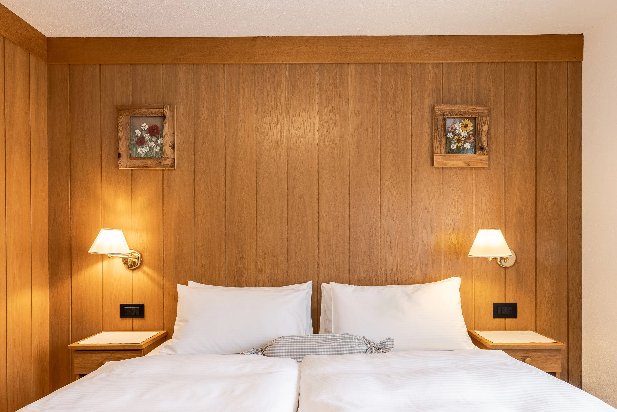 Una camera da letto con pareti di legno e due lampade da parete vicino al letto, con due quadri con fiori sopra la testata del letto.