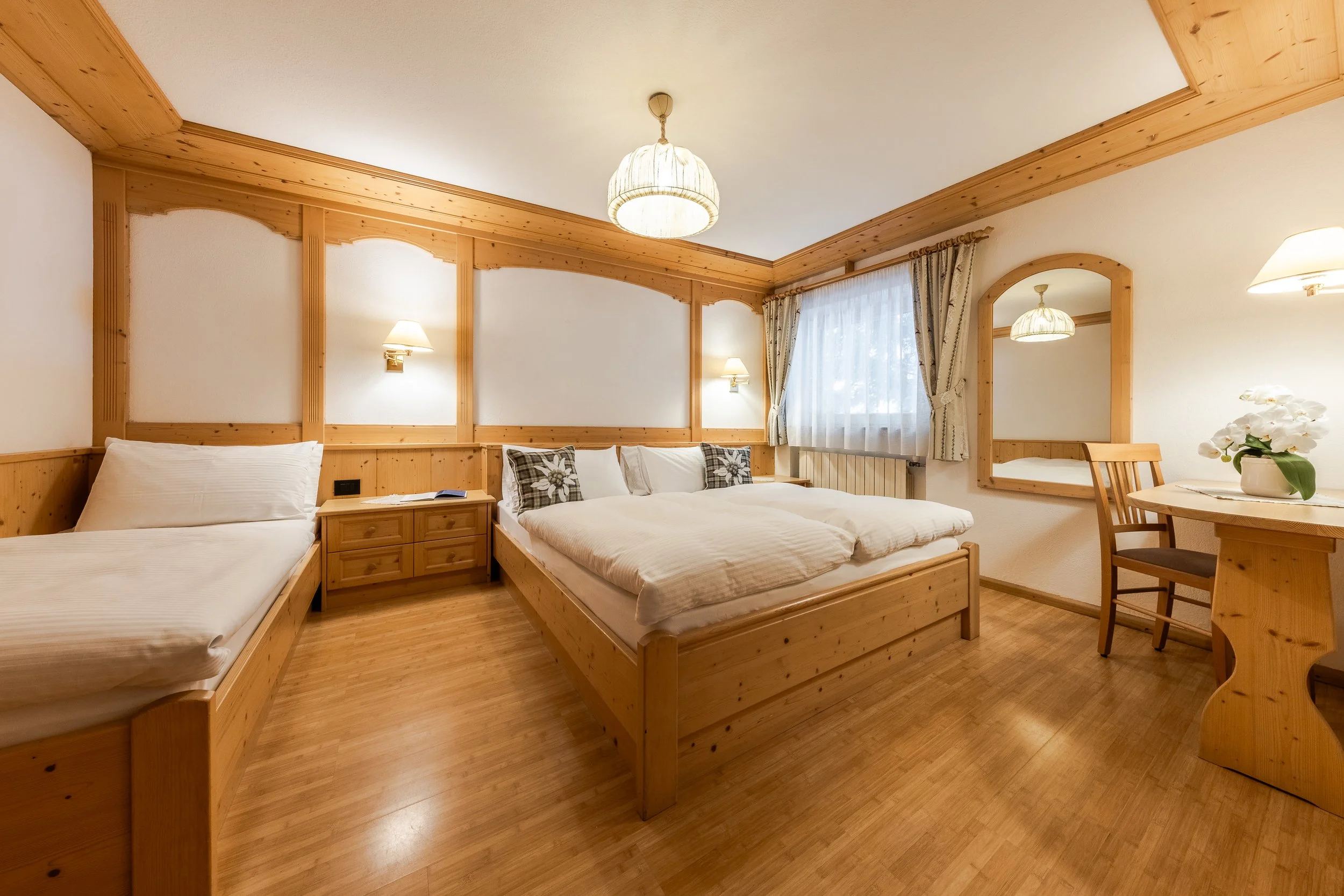 Camera con due letti con copriletto bianco, soffitto e pareti in legno, finestra con tende chiare, decorazioni floreali e specchio appeso sulla parete.