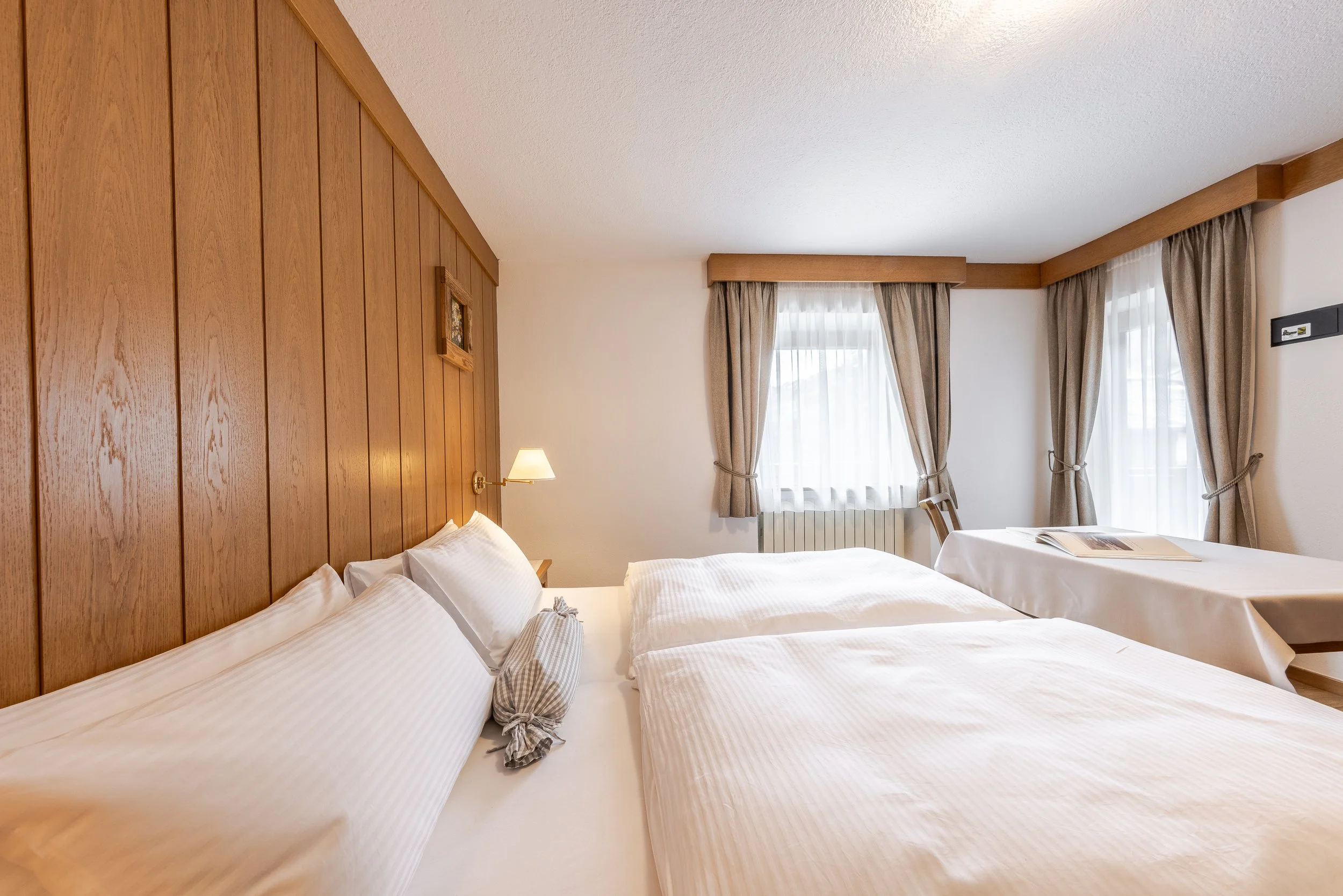 Stanza d'albergo con due letti singoli e un letto matrimoniale, pannelli di legno sul muro dietro i letti, tende beige alle finestre, lampada da parete e finestra con tende beige e colori chiari.