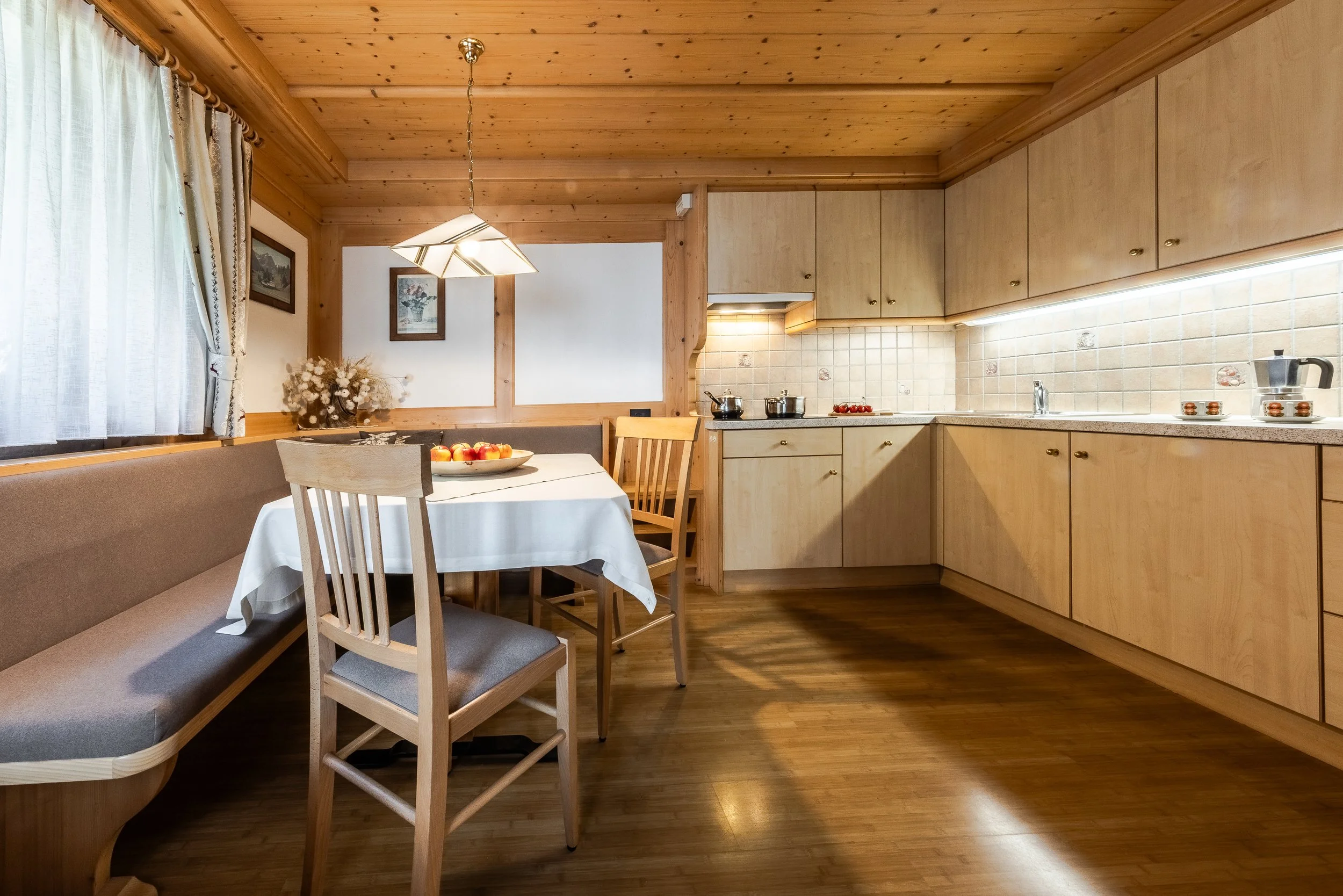 Interno di una cucina con pareti, soffitto e mobili in legno chiaro, tavolo con tovaglia bianca, piatto di mele e sedie in legno, finestre con tende leggere e quadri appesi alla parete.