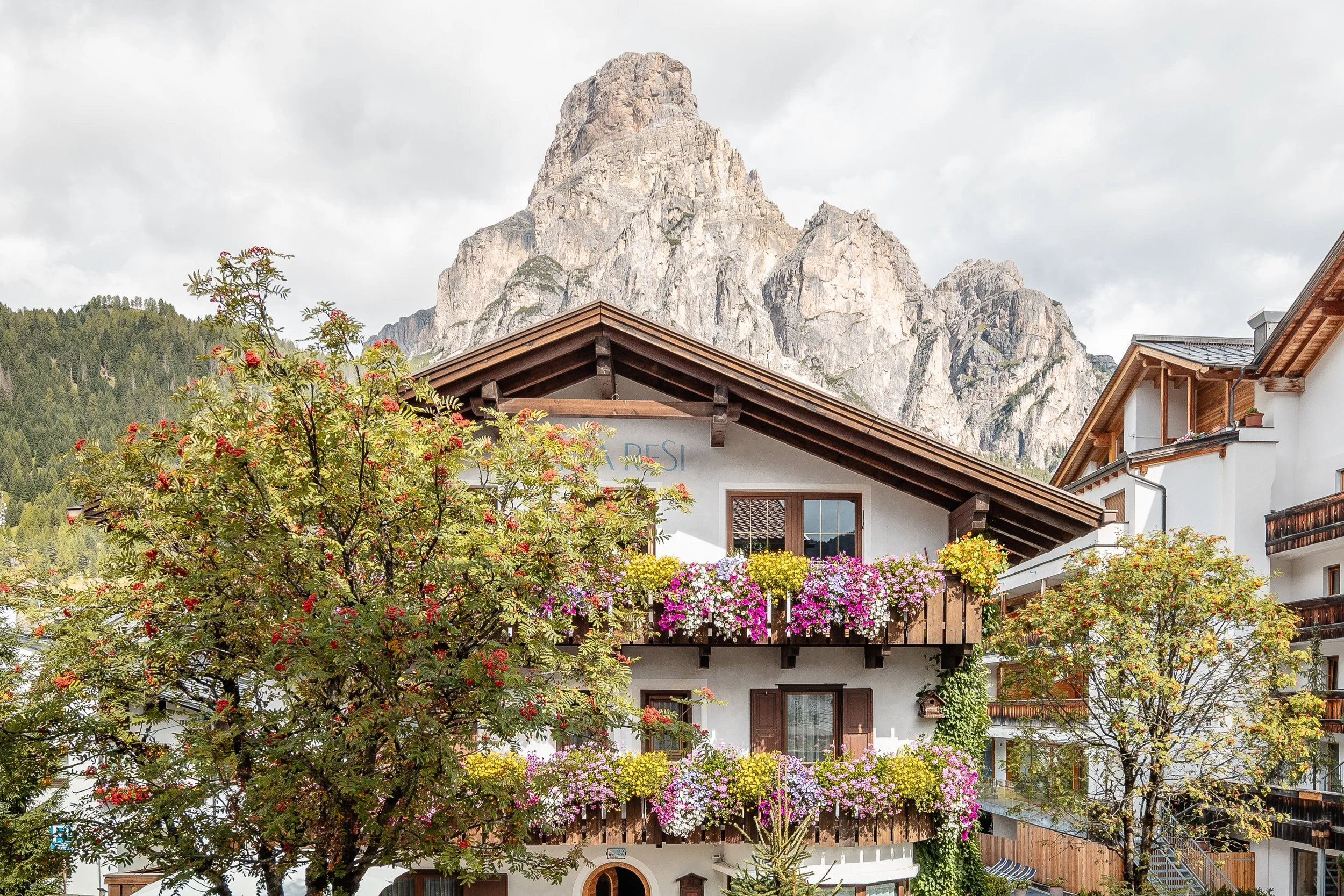 Case alpine con balconi fioriti, sullo sfondo montagne rocciose e un cielo nuvoloso.