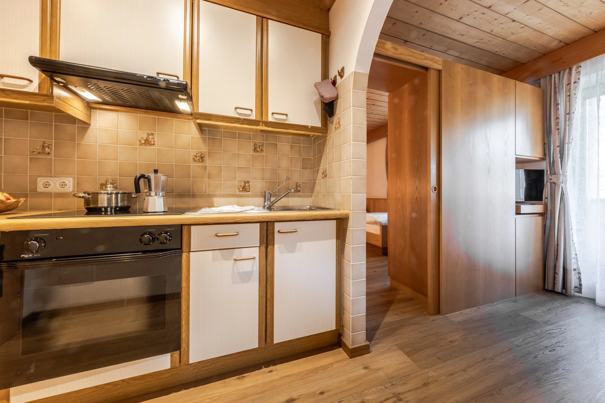 Cucina con armadietti bianchi con maniglie di ottone, piano di lavoro in legno, forno elettrico e piano cottura a gas con pentola. Dietro, pareti rivestite di piastrelle beige con decorazioni e prese di corrente, e una porta scorrevole in legno che c