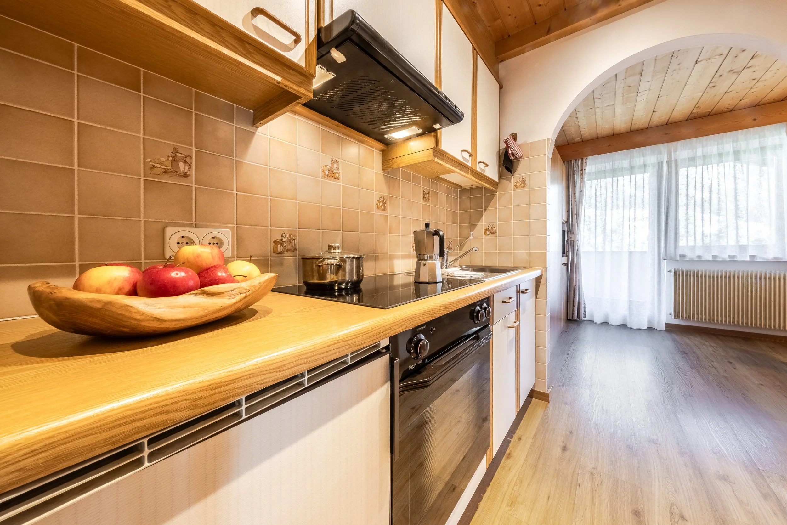 Cucina con piani in legno, pareti rivestite di piastrelle beige con decorazioni di oggetti da cucina, balcone con tende e porte scorrevoli con vista sulla natura.