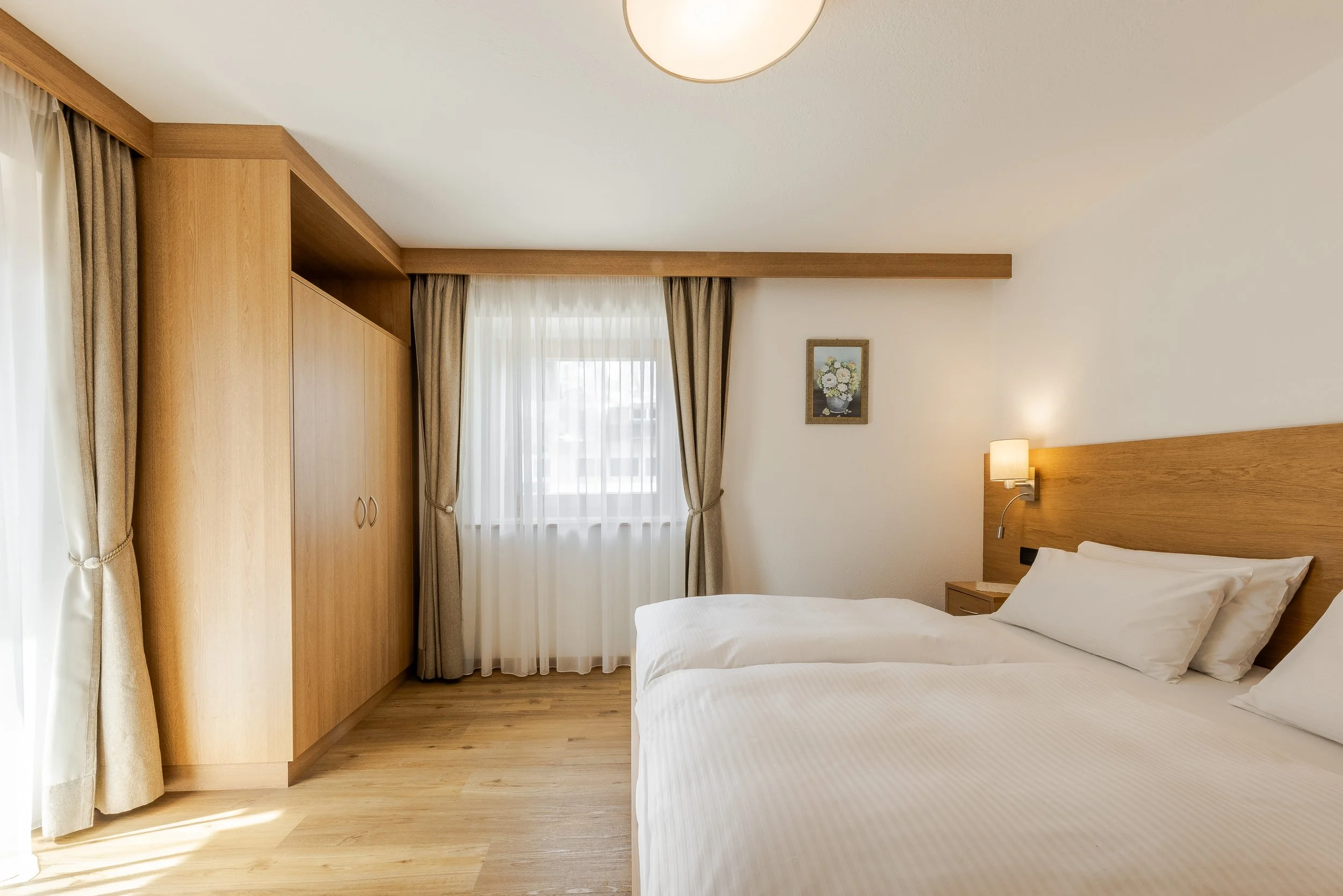 Stanza d'hotel con due letti, armadio in legno, finestra con tende beige e quadro floreale sulla parete.