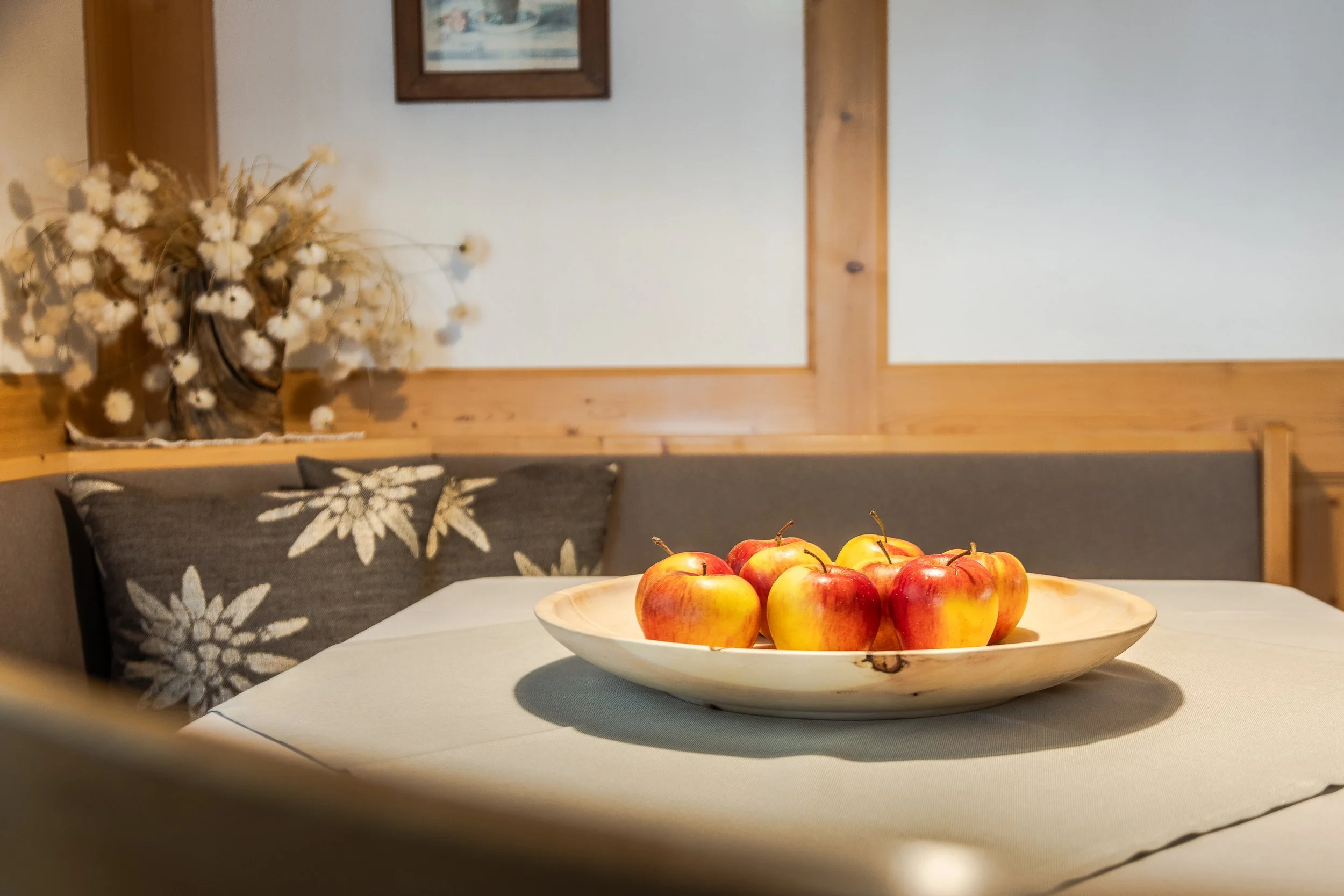 Un piatto di mele rosse e gialle su una tovaglia bianca vicino a un divano con cuscini decorativi con fiori. Sullo sfondo, pareti chiare con cornici di legno e una decorazione floreale.