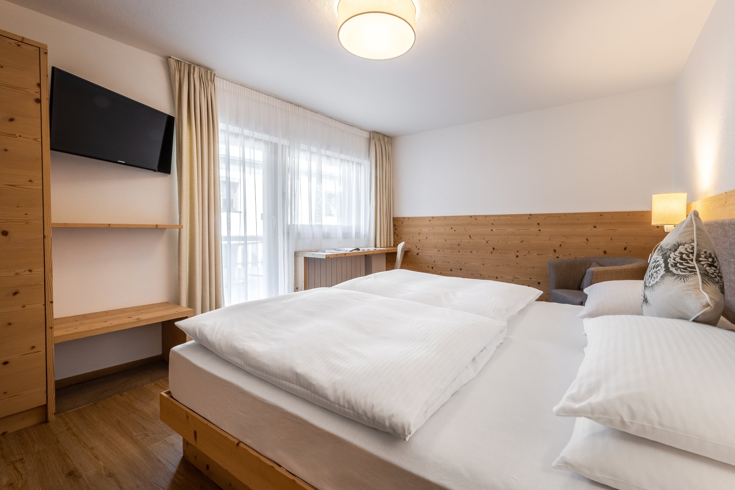 Camera da letto con due letti singoli con biancheria bianca, un comodino con lampada, una finestra grande con tende chiare, un televisore montato sulla parete, pareti bianche e pannelli di legno chiaro decorativi.
