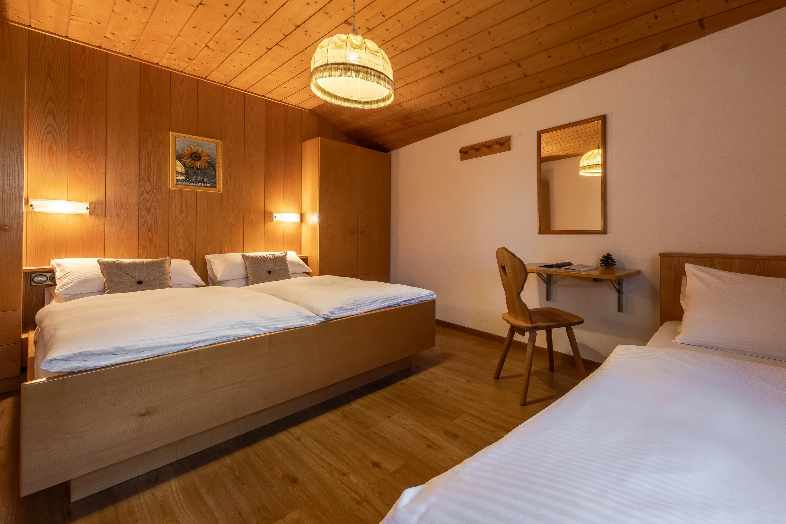 Camera con pareti e soffitto in legno, tre letti con biancheria bianca, un tavolo con sedia di legno, quadri e specchio appesi, illuminazione calda.