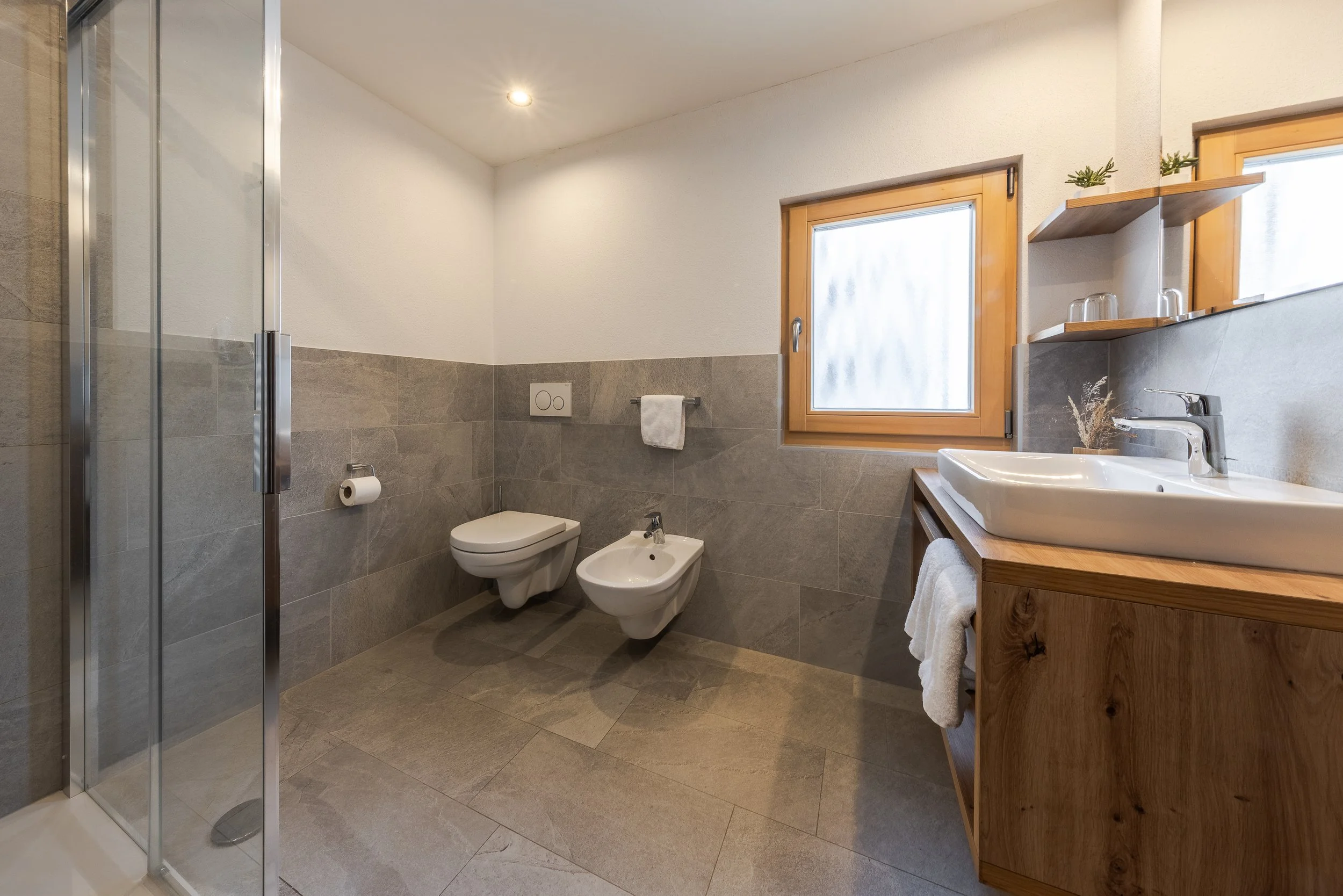 Bagno moderno con pareti e pavimenti in piastrelle di colore grigio, finestre con cornici di legno, e arredi in legno chiaro. Include una doccia con porta in vetro, un gabinetto, un bidet, e un lavabo con un mobile in legno.