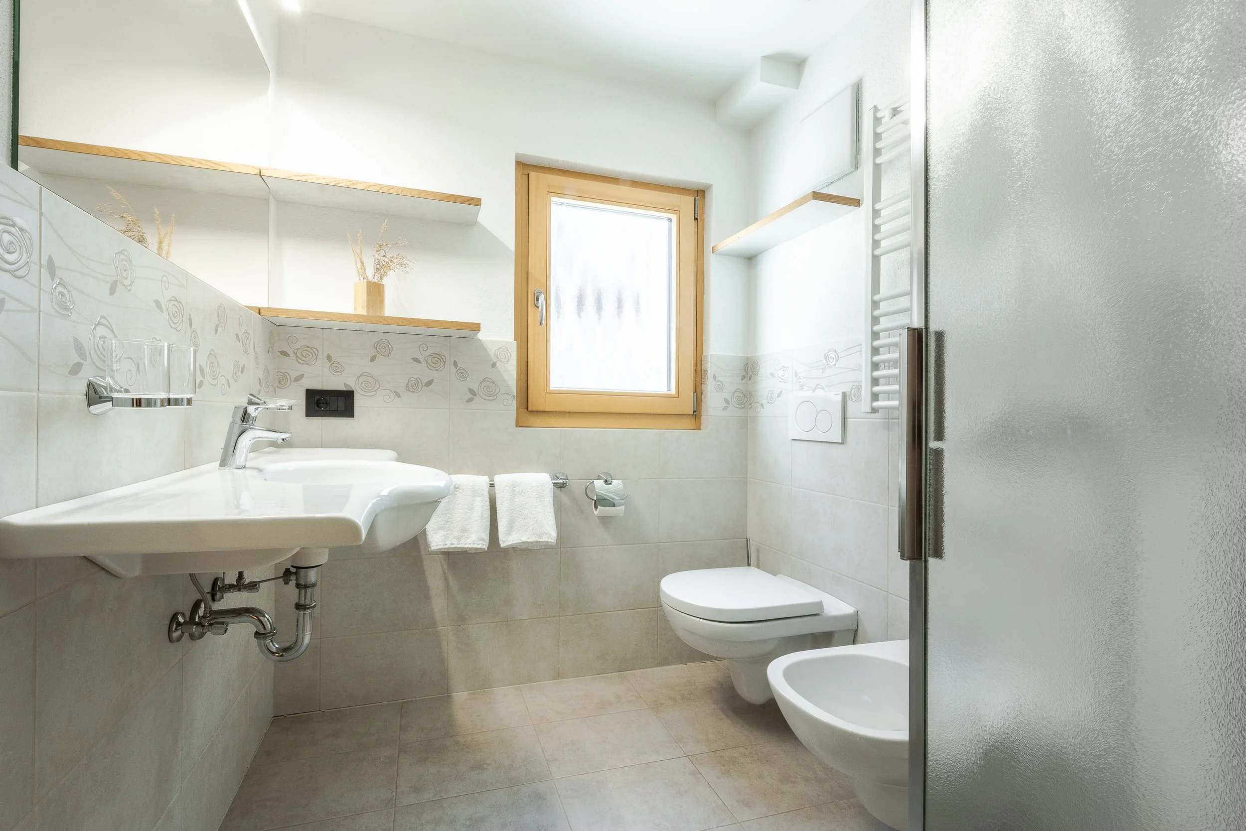 Bagno con lavabo, WC e bidet, finestra con cornice di legno, pareti e pavimenti chiari con decorazioni floreali, portasciugamani con asciugamani bianchi, radiatore a scaldasalviette, porta in vetro satinato.