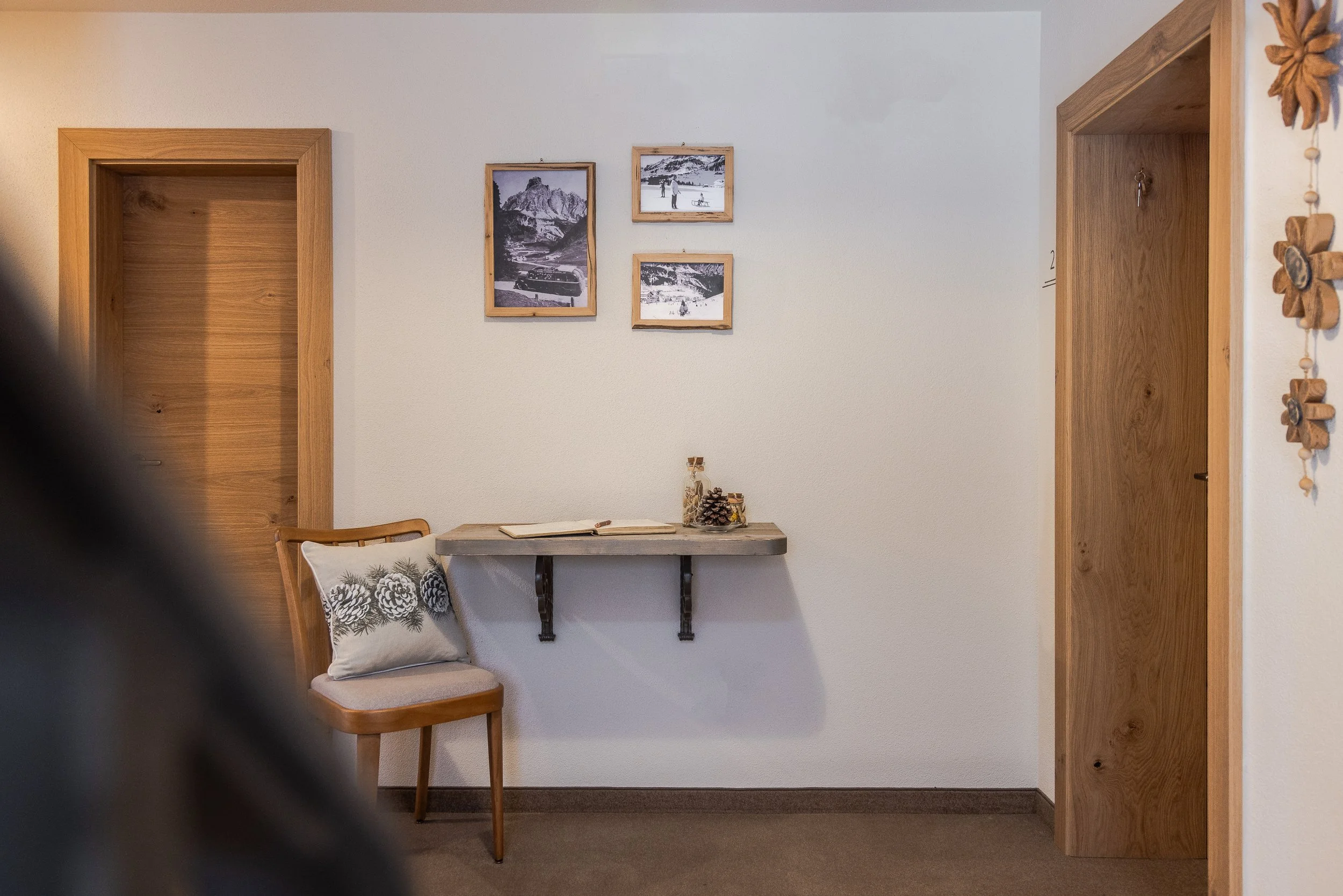 Interno di una stanza con parete bianca, tre quadri con cornici di legno e alcune foto di paesaggi invernali. In basso, un tavolo con un libro aperto, decorazioni natalizie e una sedia con cuscino con disegni di pigne.