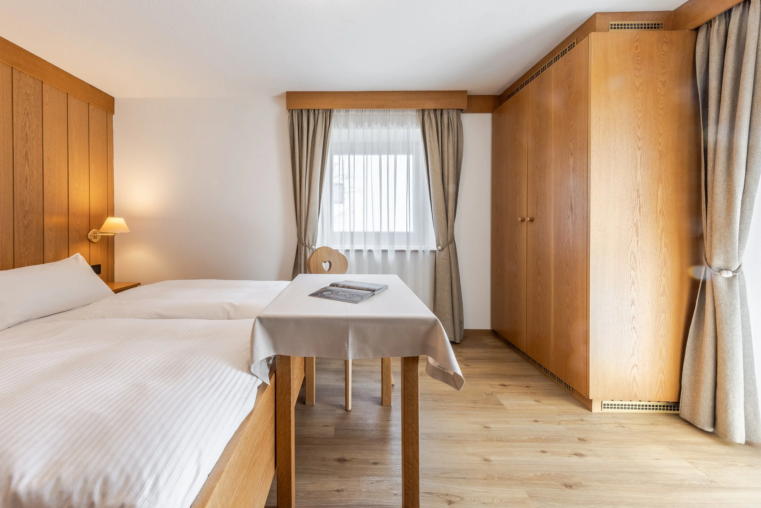 Camera di hotel con due letti a baldacchino con lenzuola bianche, pareti di legno, finestra con tende beige e luce da muro, tavolo con riviste, armadio in legno e pavimento in legno.