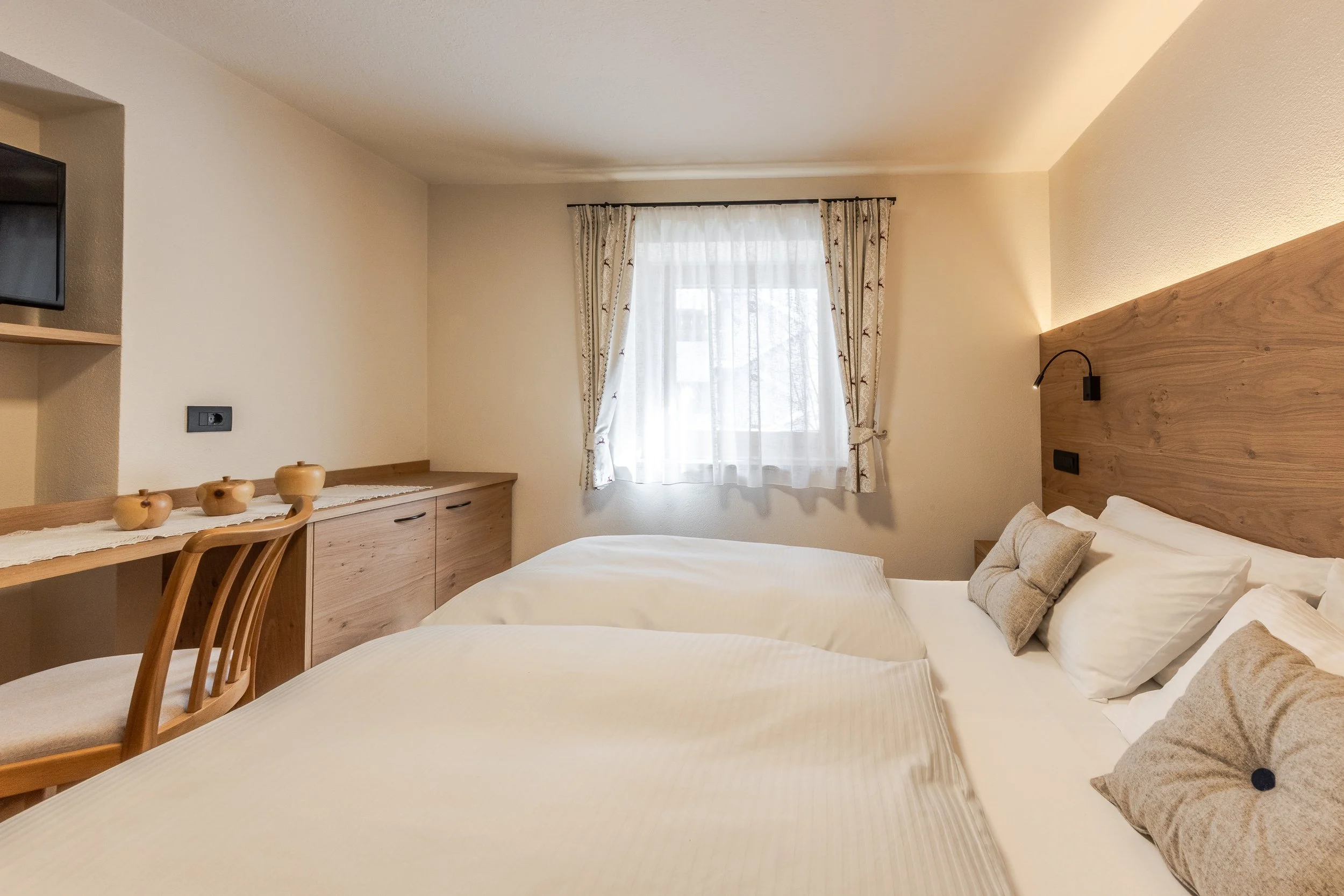Camera di hotel con letto matrimoniale, testiera in legno, cuscini beige, finestra con tende chiare, scrivania con decorazioni di ceramica e televisore.