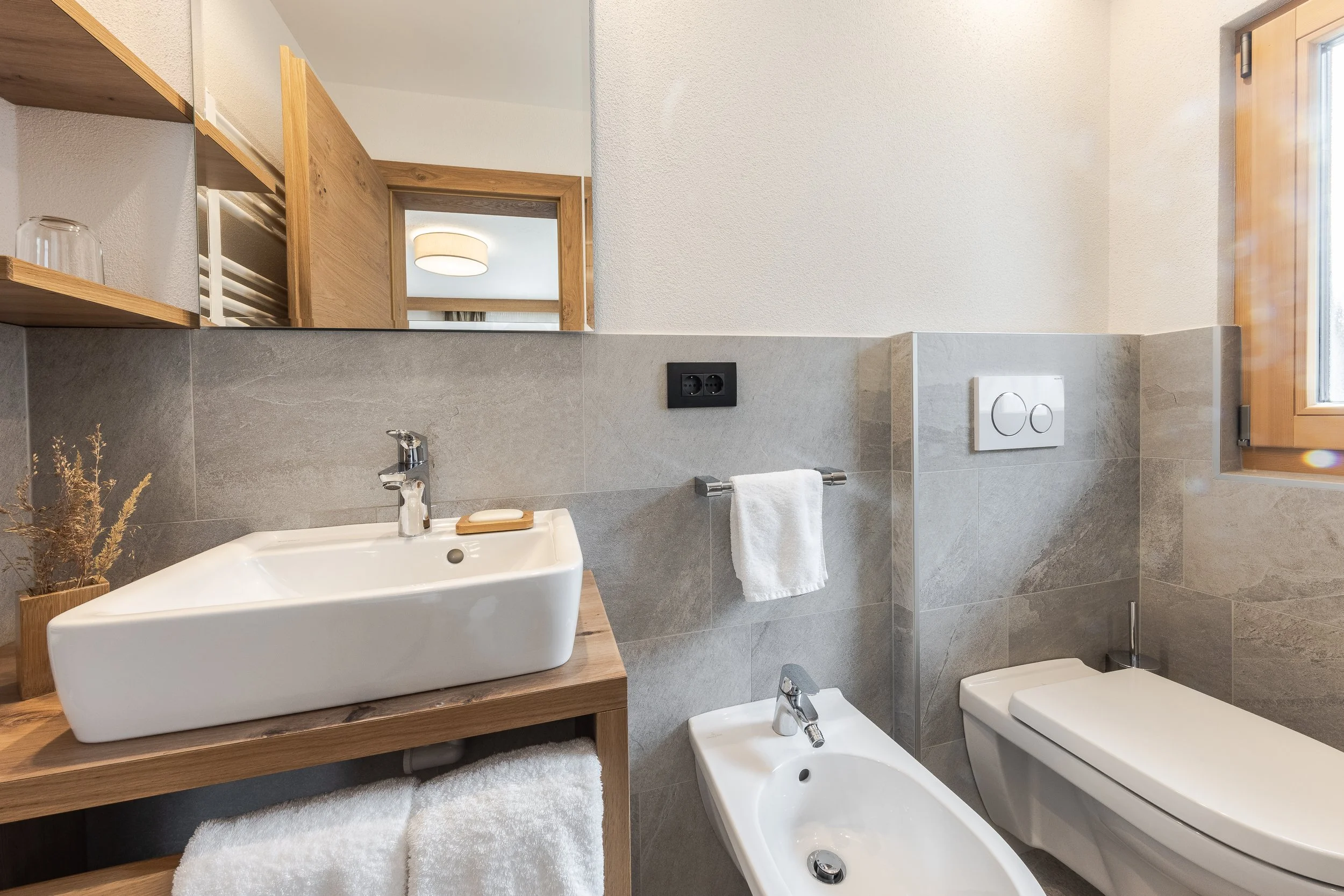 Interno di un bagno moderno con lavabo, bidet, WC, grande specchio, pareti rivestite in piastrelle grigie e decorazioni in legno, e finestra con vetri trasparenti.