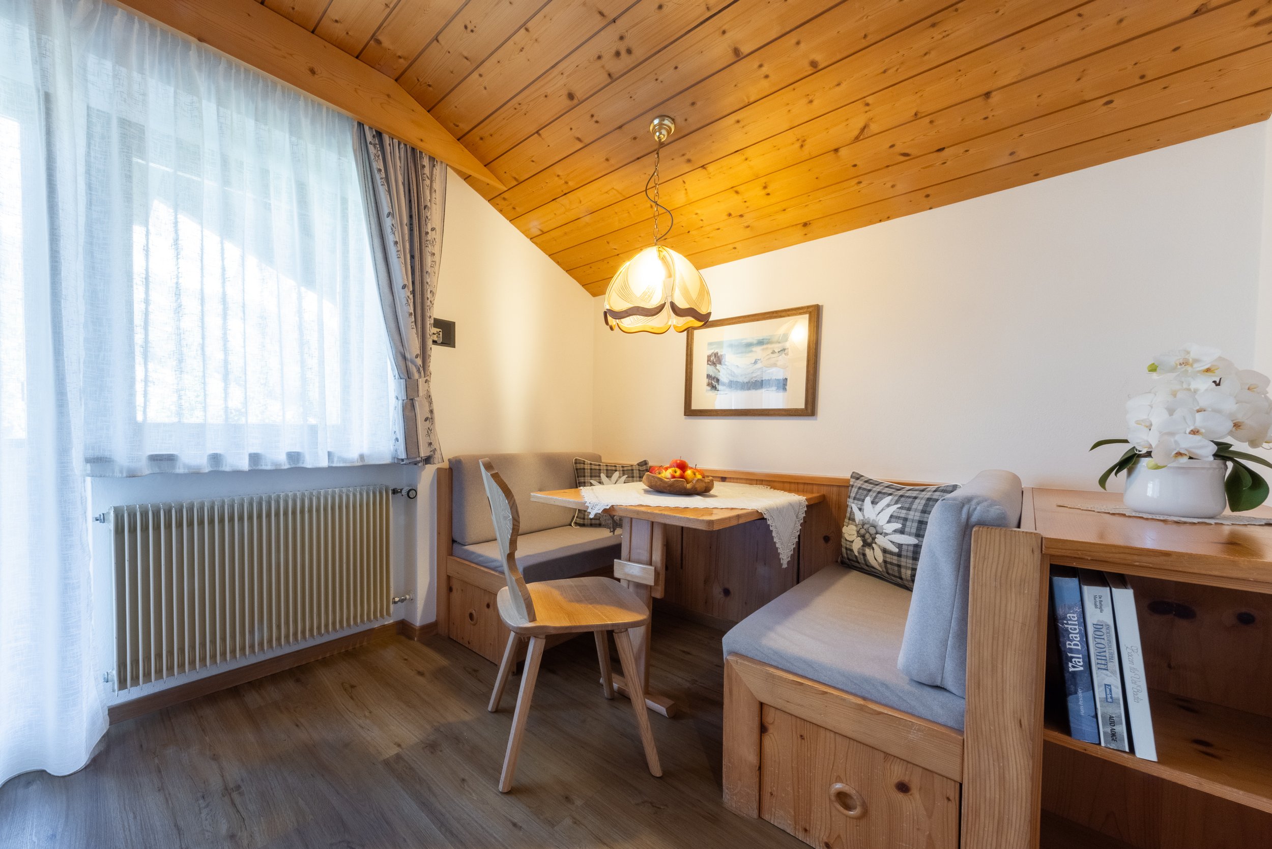 Interno di una stanza in stile rustico con soffitto in legno, una lampada appesa, un quadro appeso alla parete, una finestra con tende di tessuto trasparente, un divano con cuscini a quadri, un tavolo con una ciotola di mele e alcune sedie in legno, 