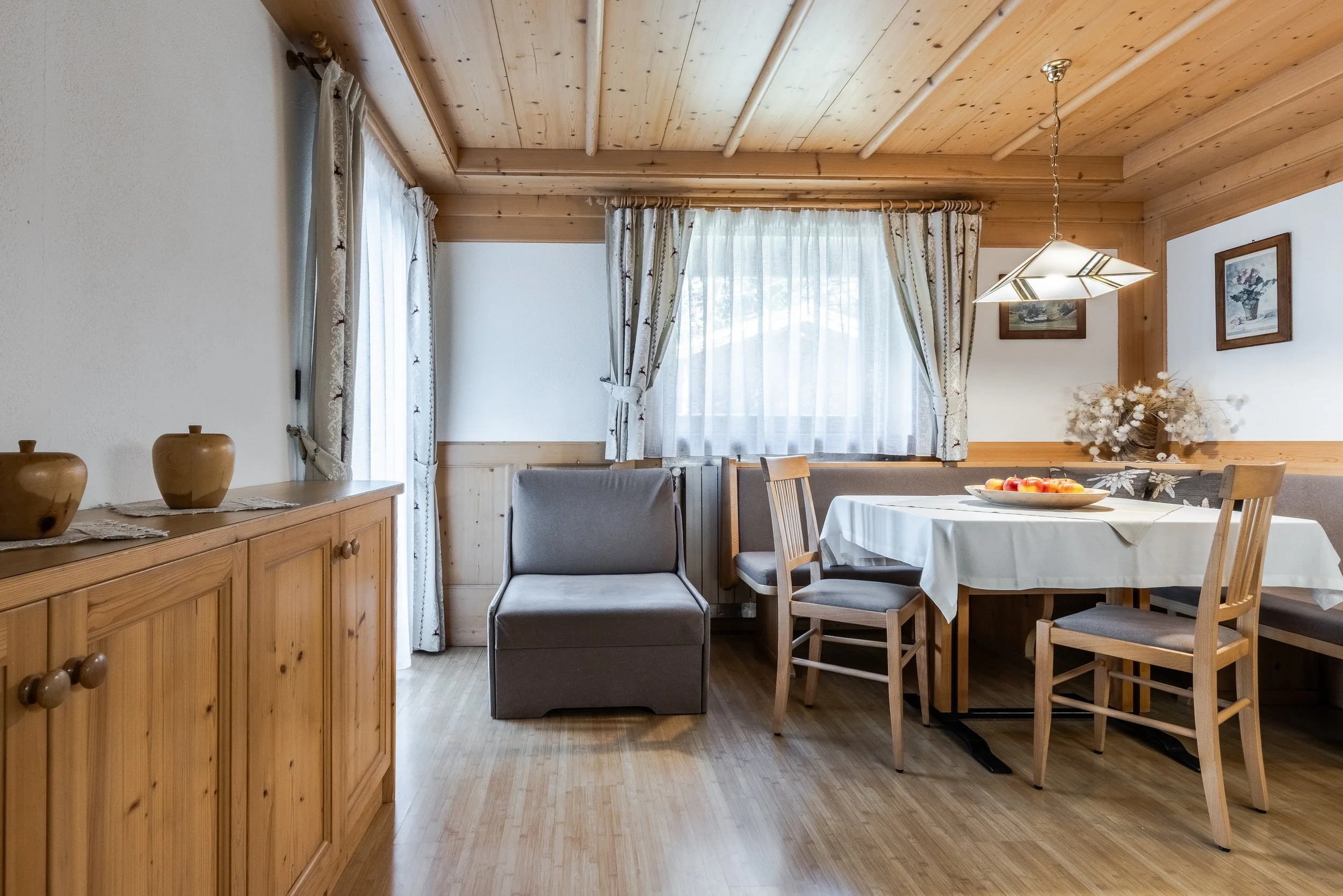Interno di una cucina o soggiorno con pareti di legno, grande finestra con tende bianche, tavolo rotondo con tovaglia bianca, sedie di legno, un divano grigio, un mobile in legno con vasi decorativi, quadri alle pareti e una pianta con fiori secchi.