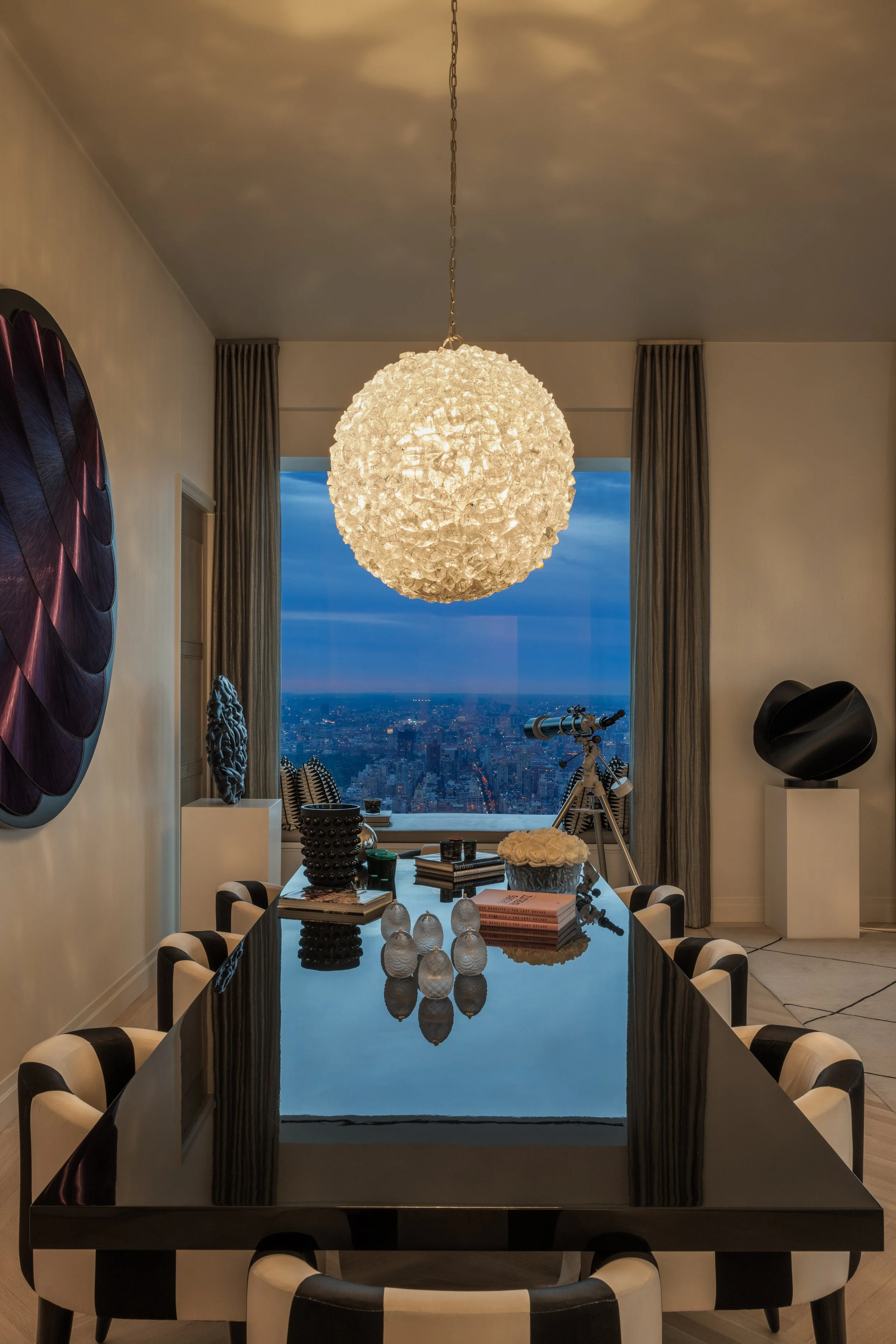 Modern dining room with black glass table, black and white chairs, large spherical pendant light, and city view through floor-to-ceiling windows, with abstract black and purple wall art.