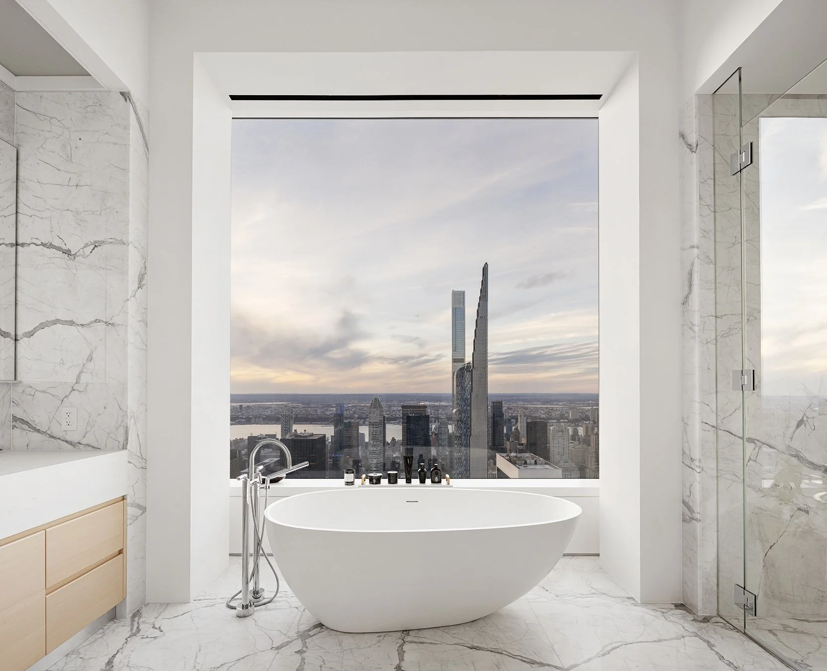 Modern bathroom with a freestanding bathtub, marble walls and floors, a large window showing a city skyline, and a glass shower enclosure.