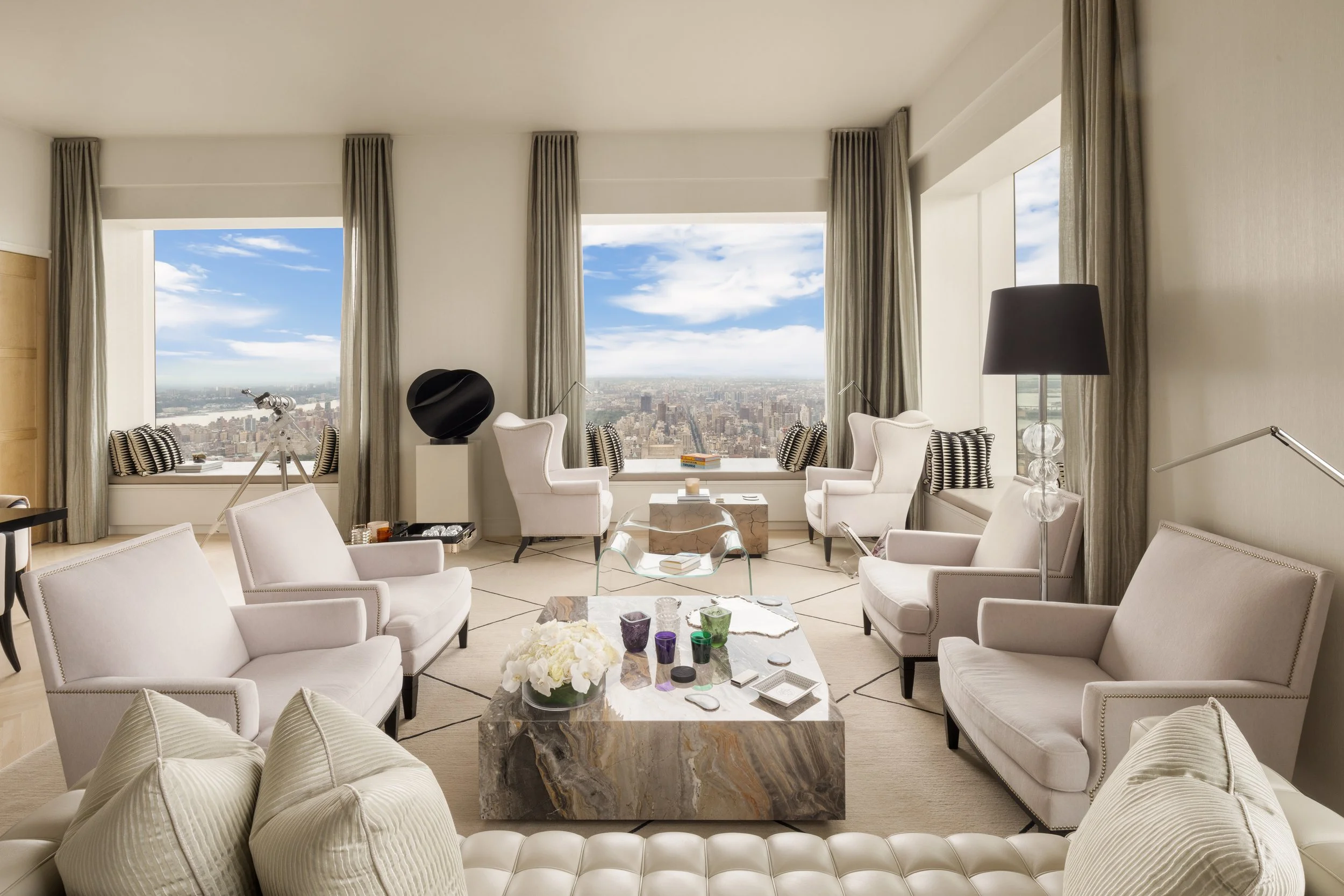 Luxurious living room with large windows showcasing city skyline, white furniture including sofas and armchairs, marble coffee table with decorative items, and a telescope near the window.
