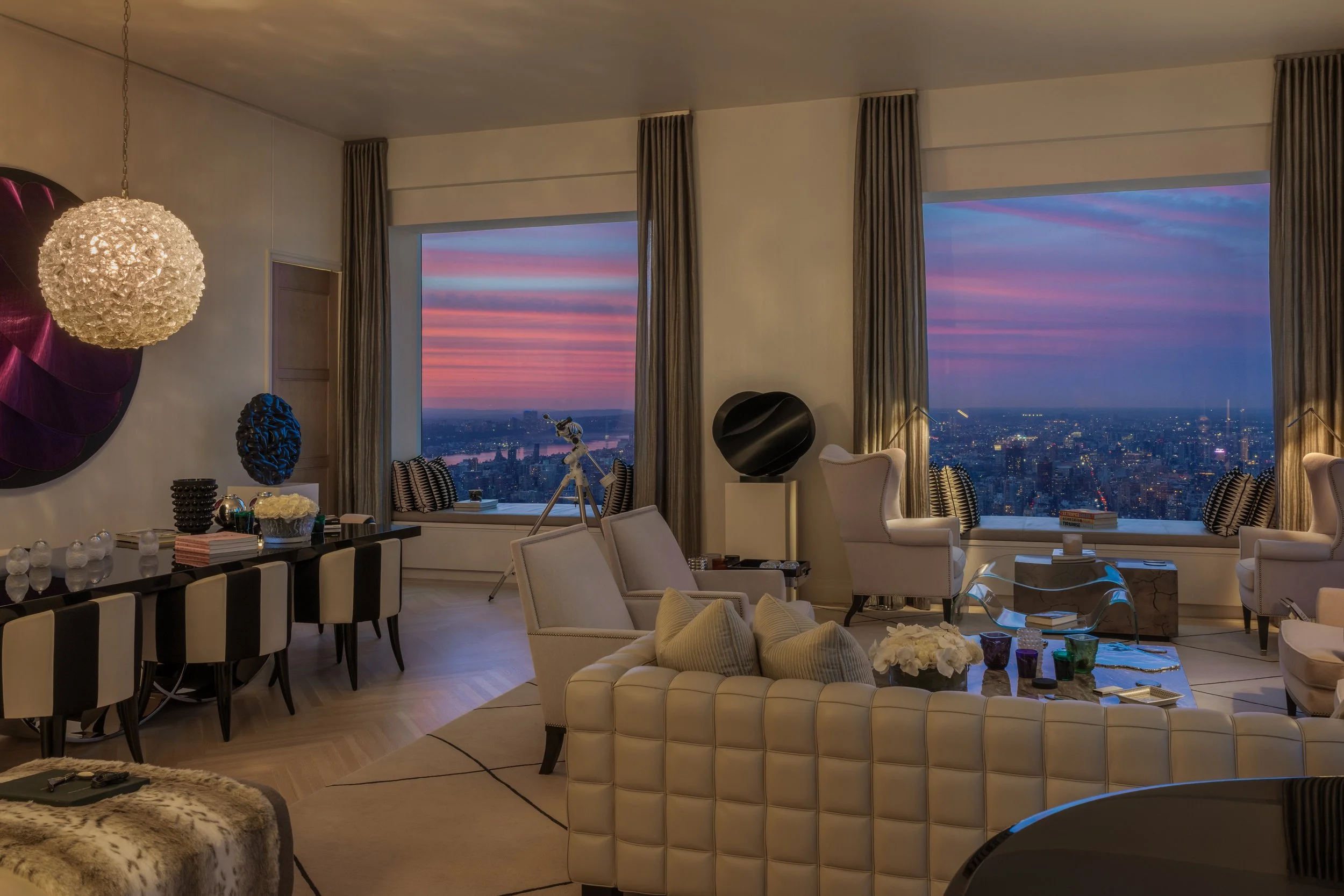 Luxurious living room with large windows showing a city skyline at sunset, furnished with white sofas, armchairs, a glass coffee table, and decorative art pieces.