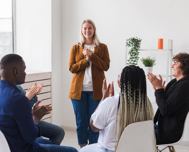 A woman leading a support group or workshop, smiling with clasped hands, surrounded by diverse participants clapping in a bright, minimalist room with a plant shelf.