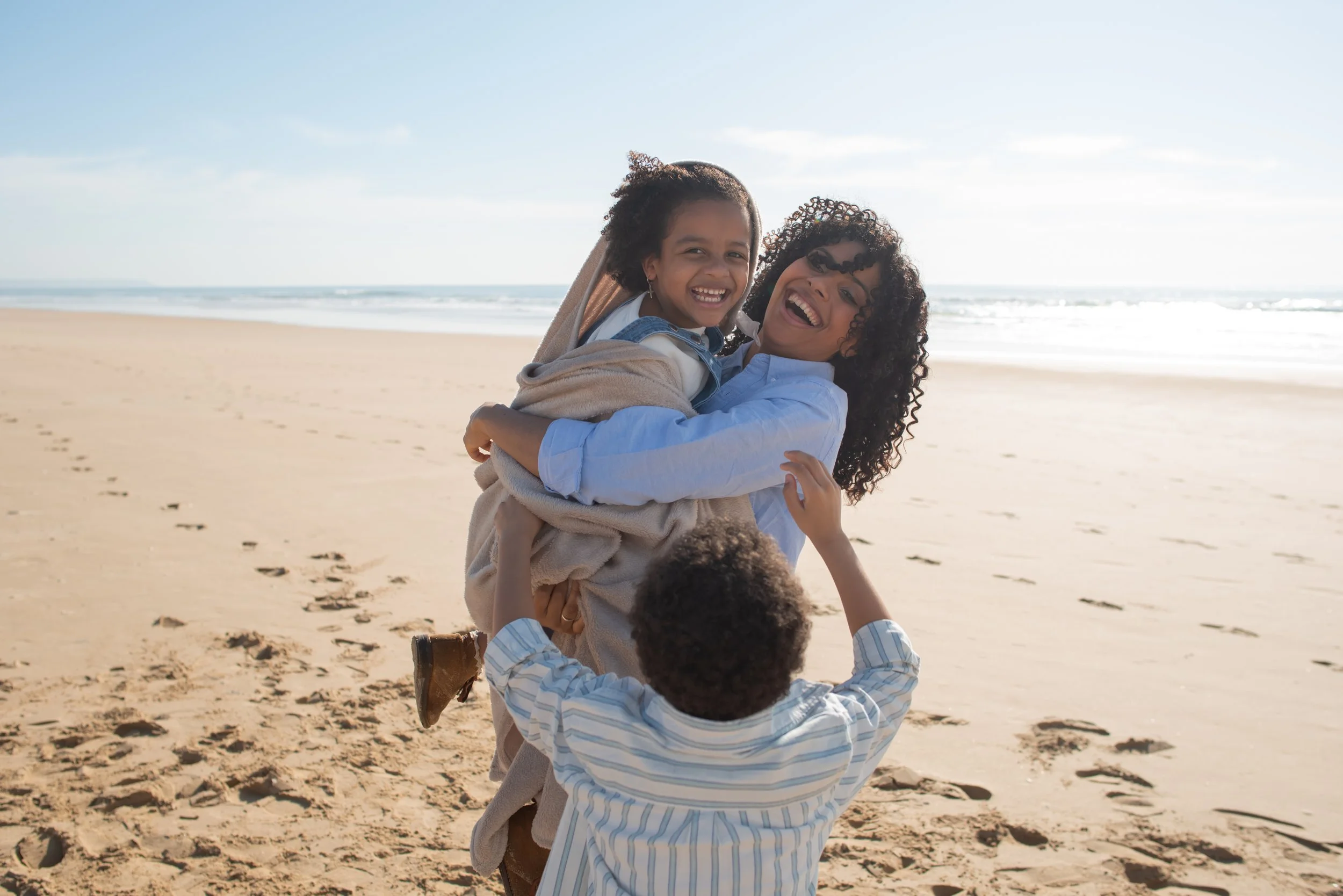 A woman is holding a girl in her arms on the beach, while a boy reaches up to play with them. The group is smiling and having fun near the ocean, with sand and footprints visible on the beach and waves in the background.