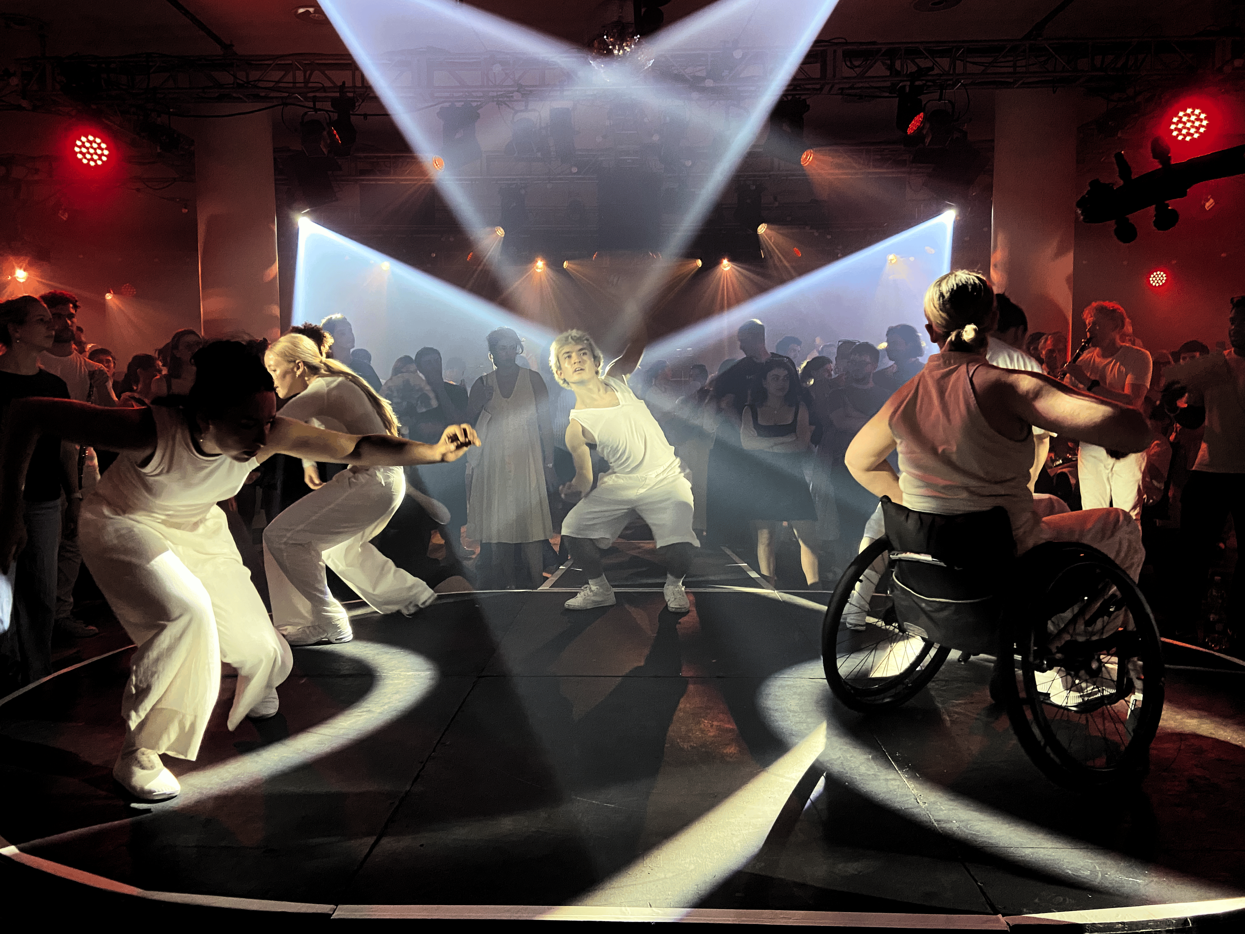 Dance performance with dancers on stage; one in a wheelchair; dramatic lighting with spotlights and audience watching.