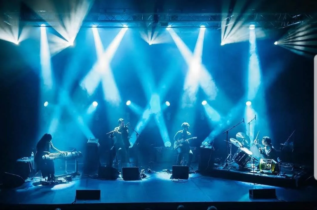 A band performing on stage with blue and white stage lighting, including musicians playing keyboards, violin, guitar, drums, and other equipment.